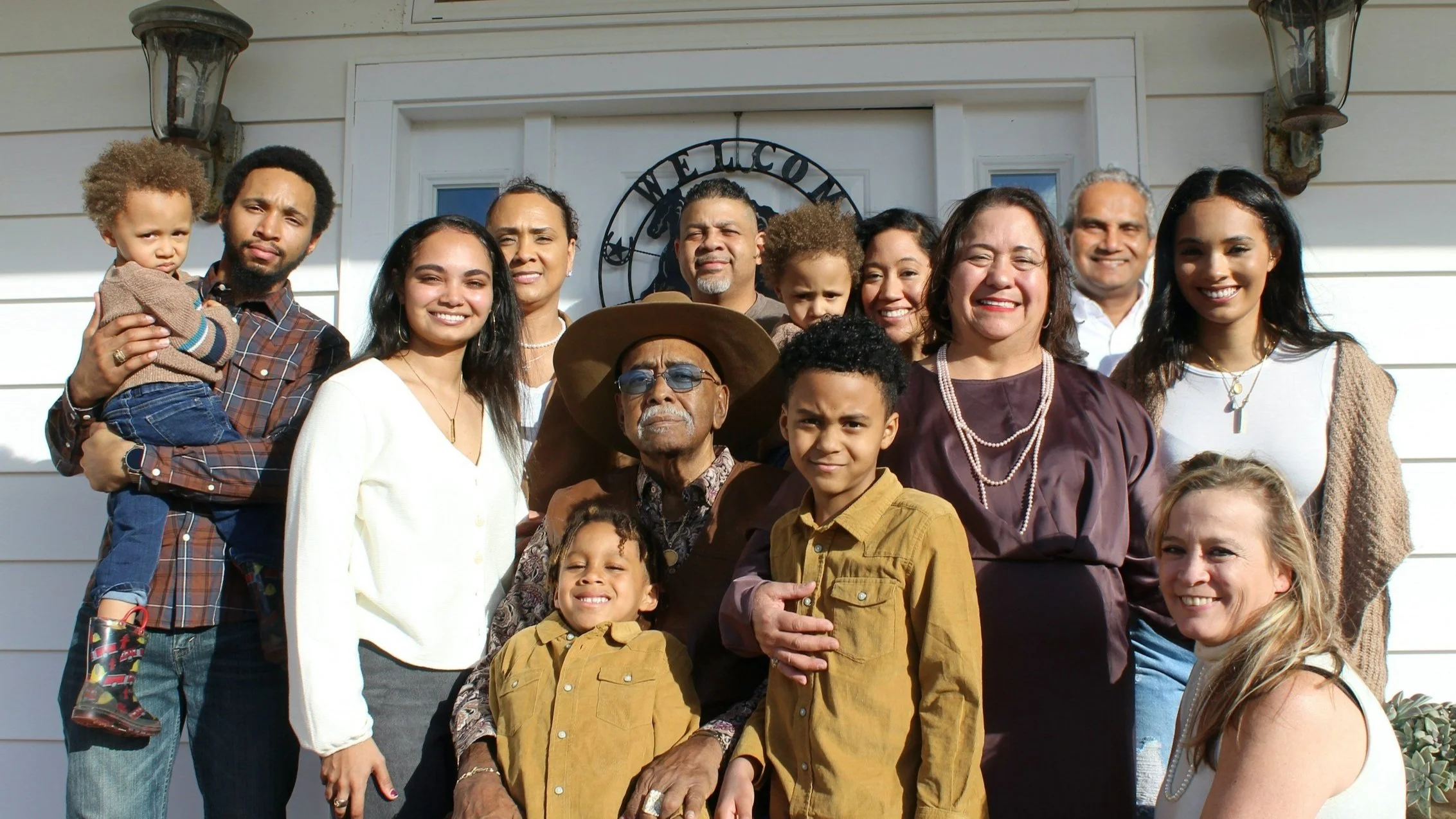 A diverse group of people, including children and adults, standing together outside a house with a welcoming sign, smiling for a group photo.