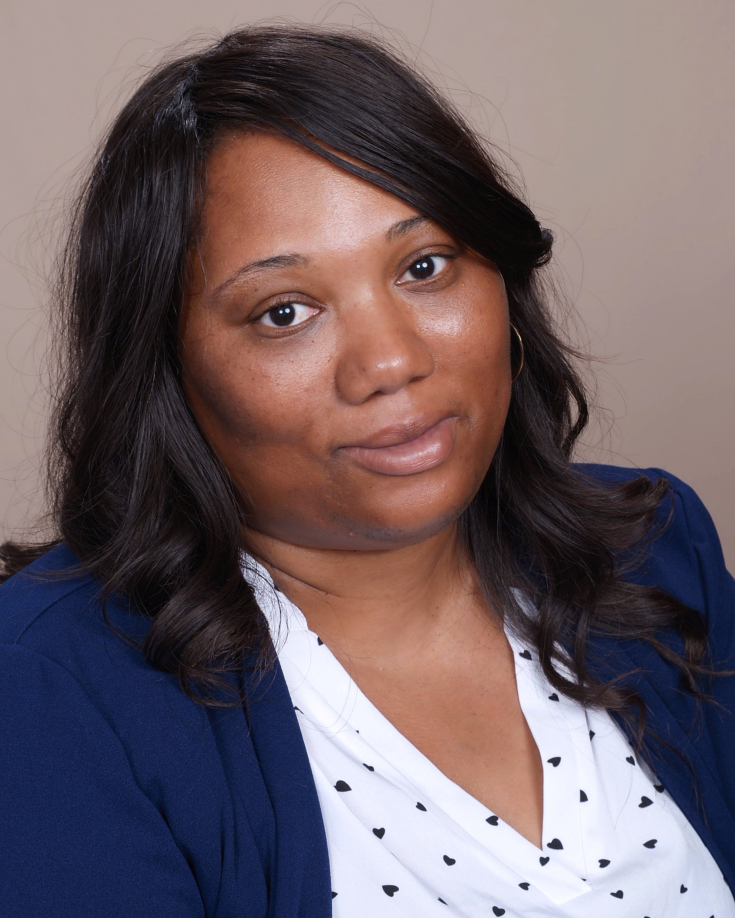 A woman with dark hair styled in loose waves, wearing a navy blue blazer over a white blouse with small black heart patterns, looking at the camera with a neutral expression.