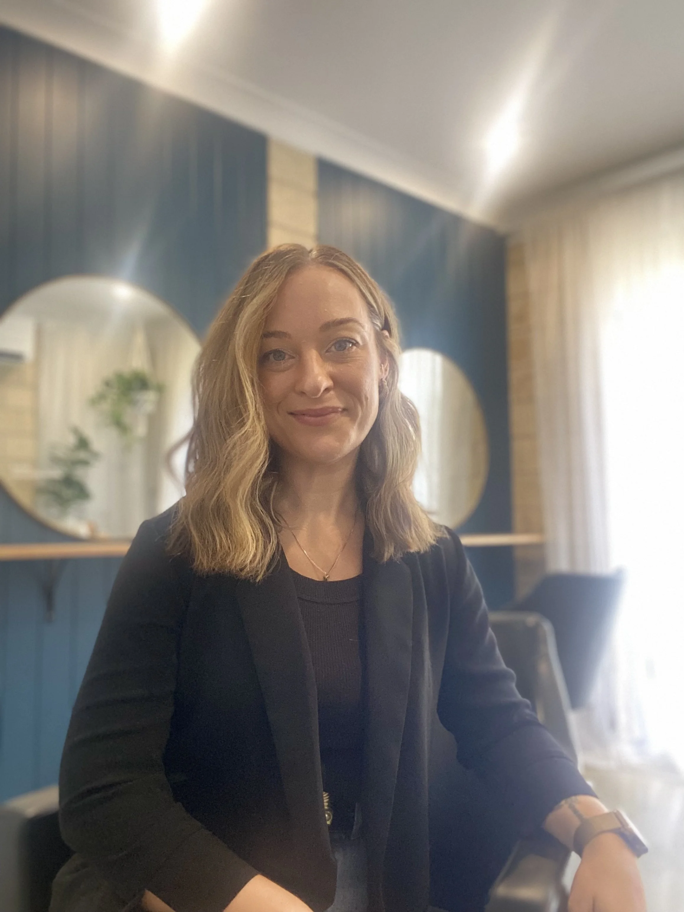 A woman with shoulder-length wavy blonde hair sitting in a room with blue walls, round mirrors, and a window with curtains, wearing a black blazer and a black top.