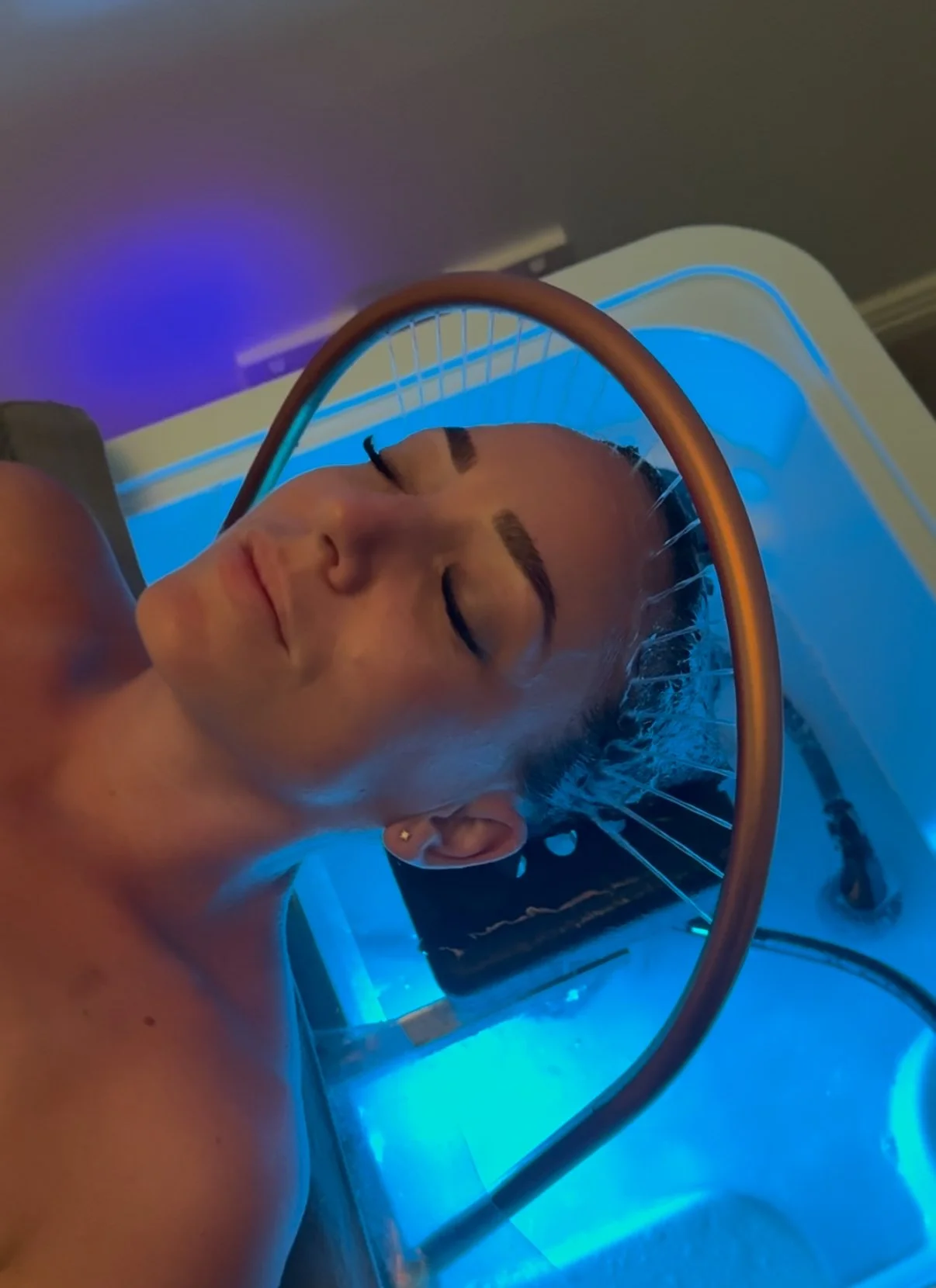 A woman undergoing a cryotherapy treatment, lying on a bed with her eyes closed, surrounded by blue lighting.