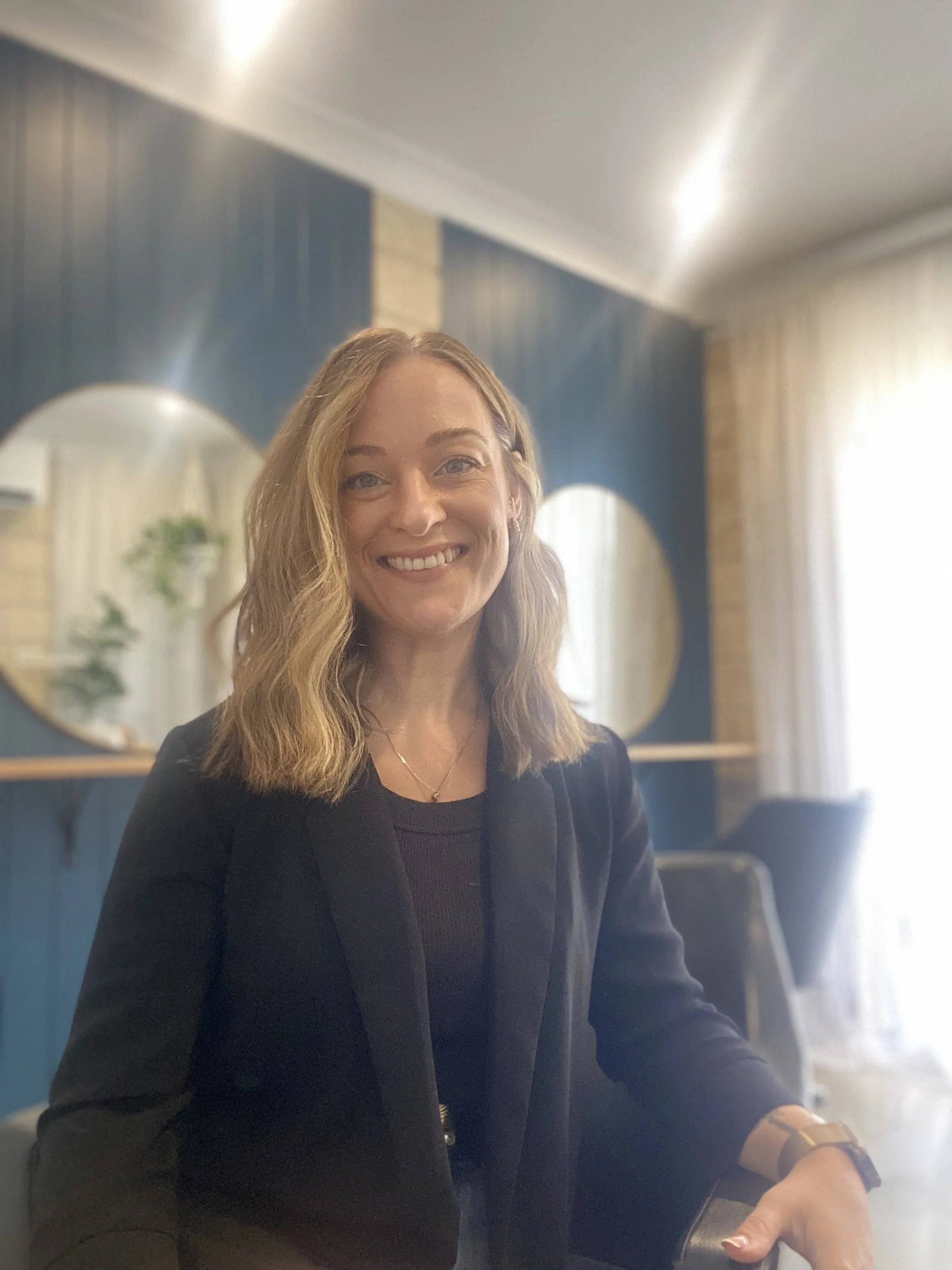 A woman with shoulder-length blonde hair smiling at the camera, wearing a black blazer and sitting indoors with a modern decor background, including two circular mirrors and blue walls.