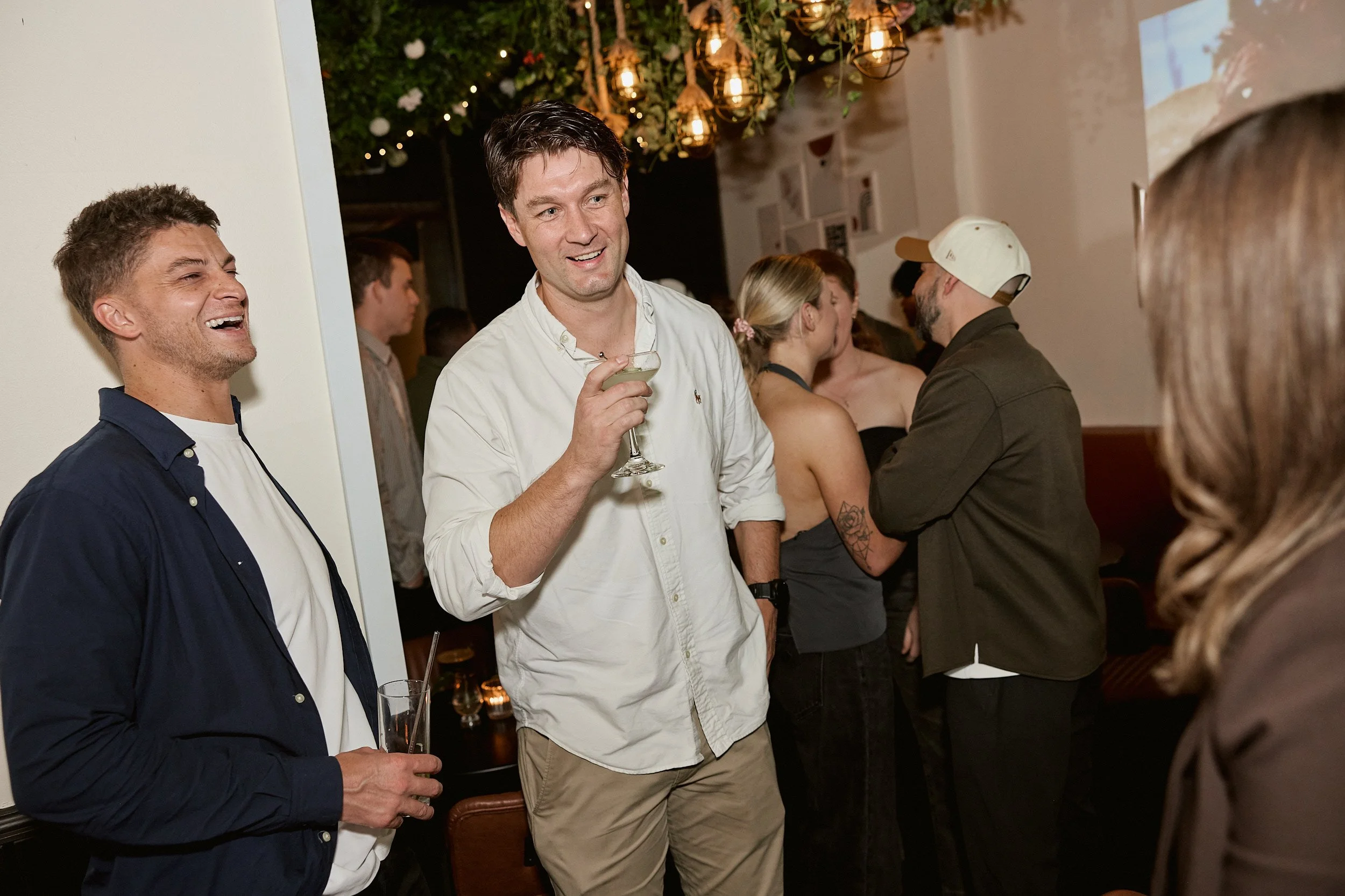 Group of people at a social gathering, talking and laughing, in a warmly lit indoor setting with decorative hanging lights and greenery.
