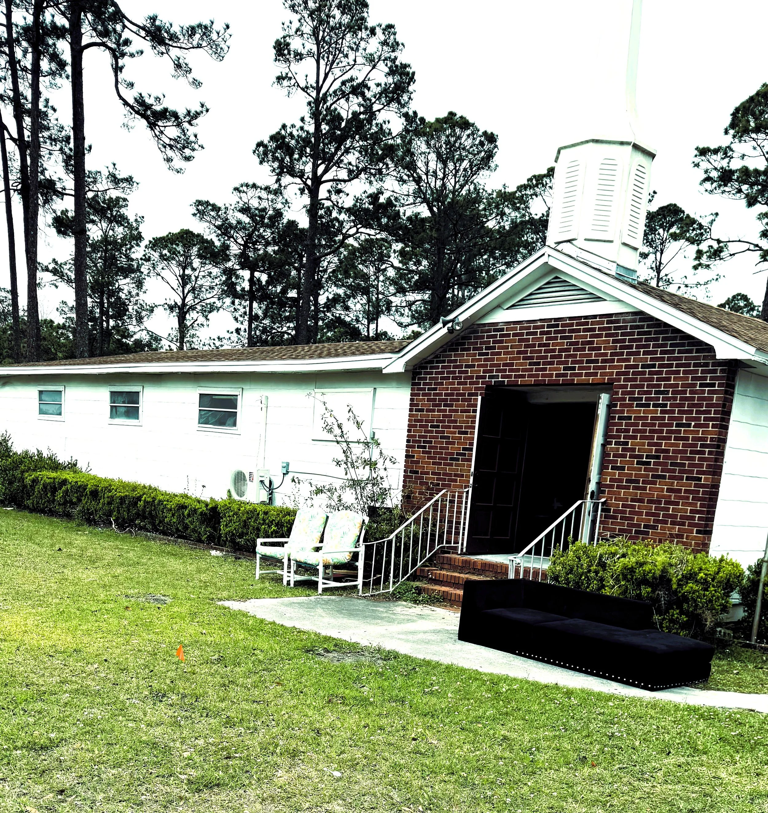 A white and red brick building with a small chimney, surrounded by green grass, bushes, and tall trees in the background. There are white chairs, a black couch, and a small ramp leading to the entrance.
