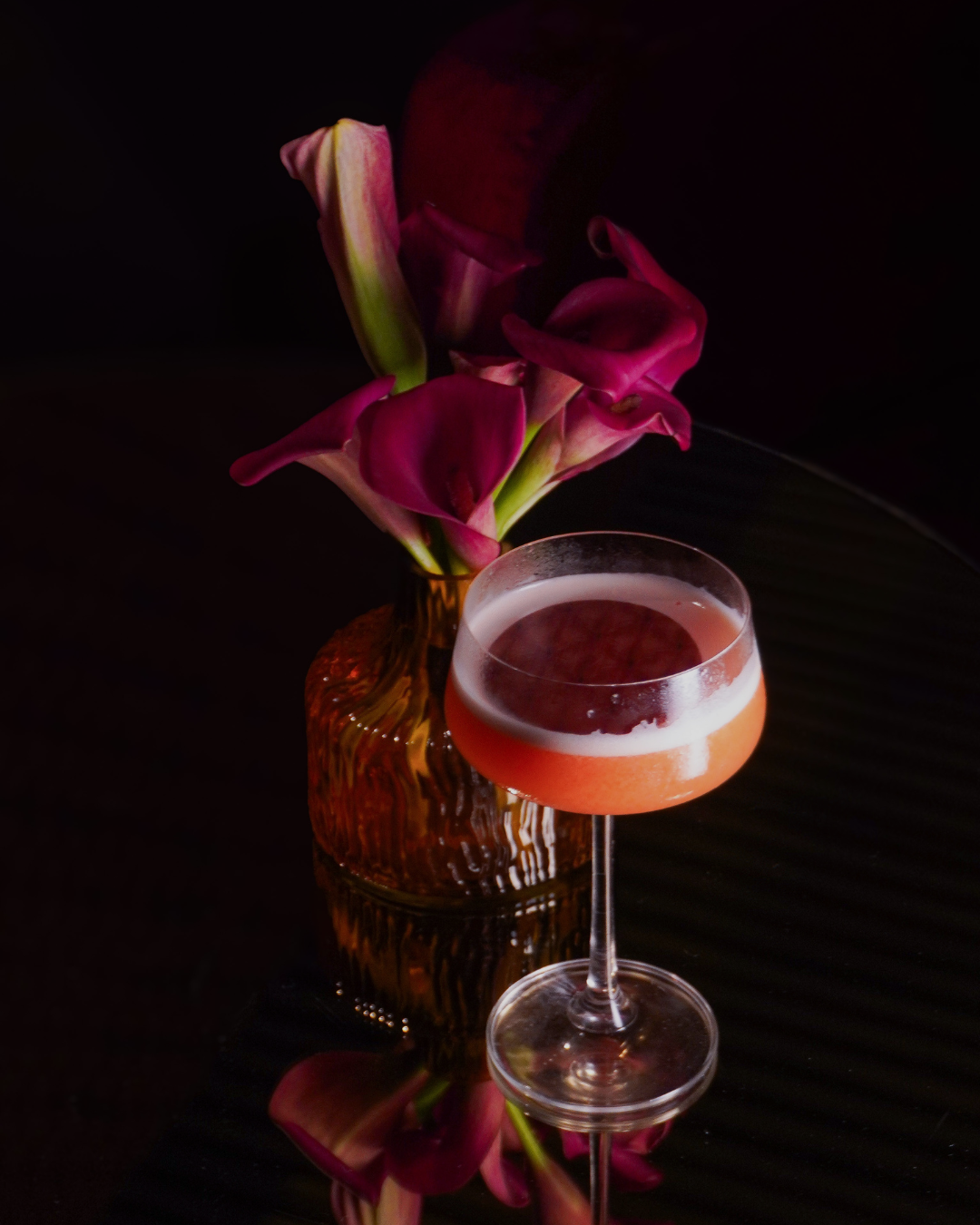 Vaso de flores rosas e uma taça de drink com cor laranja em primeiro plano, com fundo escuro.