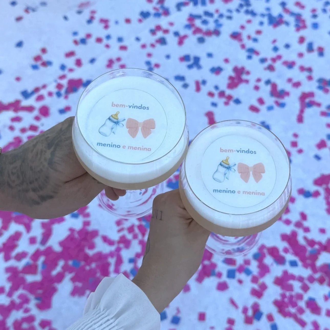 Duas mãos segurando copos de papel decorados com um desenho de mamadeira e coração, escrito 'bem-vindos, menino e menina'; ao fundo, confetes rosa, azul e branco.