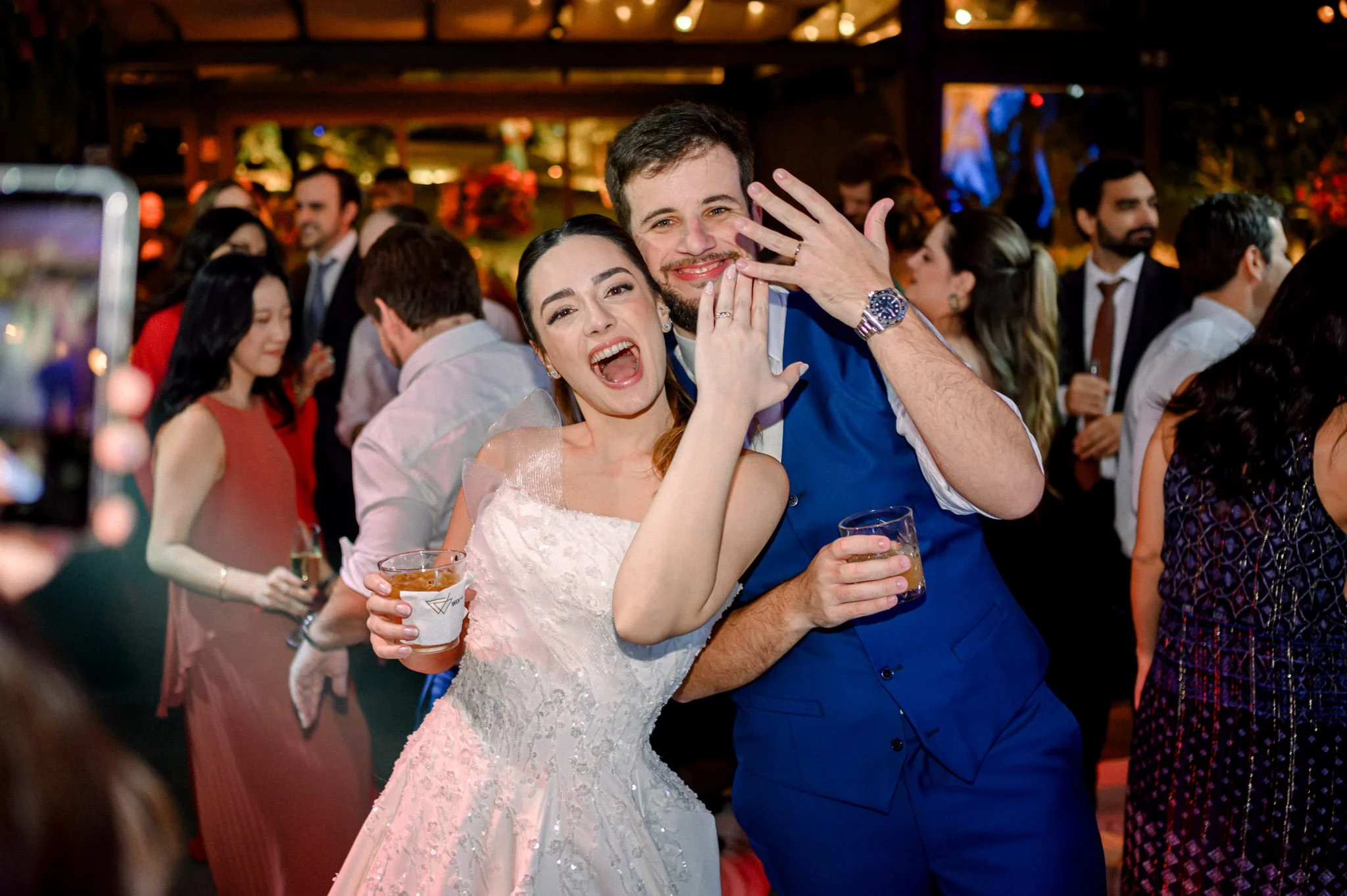 Casal sorridente brindando e celebrando em uma festa de casamento com convidados ao fundo.