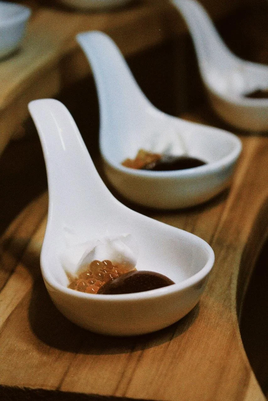 Três porções de caviar e molho de soja em tigelas brancas com formato diferenciado, dispostas sobre uma tábua de madeira.