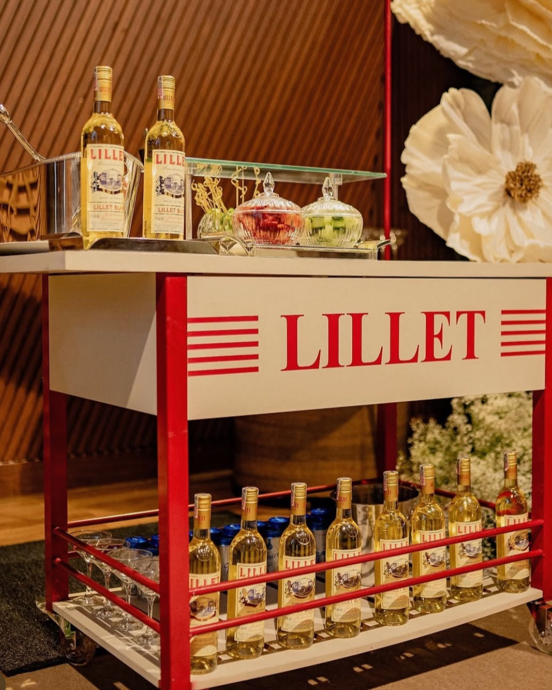 Carrinho com garrafas de licor Lillet, alguns copos e potes de doces, decorado com flores brancas ao fundo.