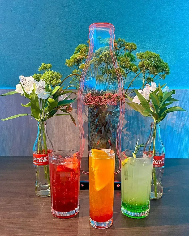 Três copos de coquetel coloridos com gelo, uma garrafa de vidro com flores brancas, uma placa de néon em forma de garrafa de Coca-Cola, e duas garrafas de vidro com flores, tudo sobre uma mesa de madeira.