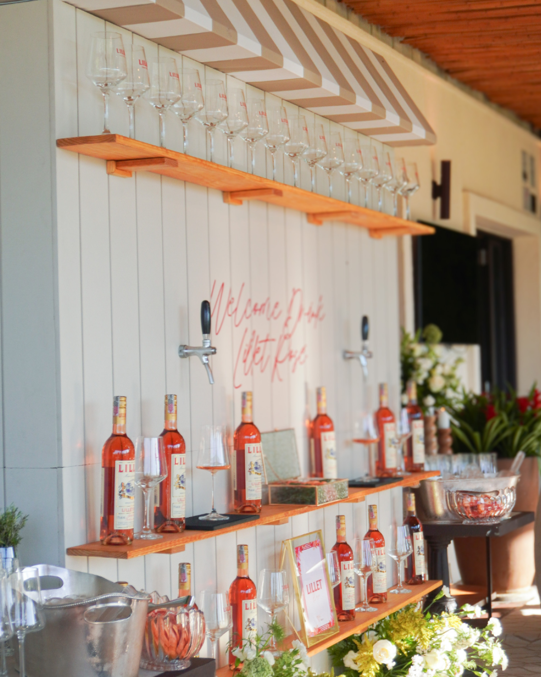 Display com garrafas de rosé, taças de vinho, e decoração floral em um ambiente ao ar livre