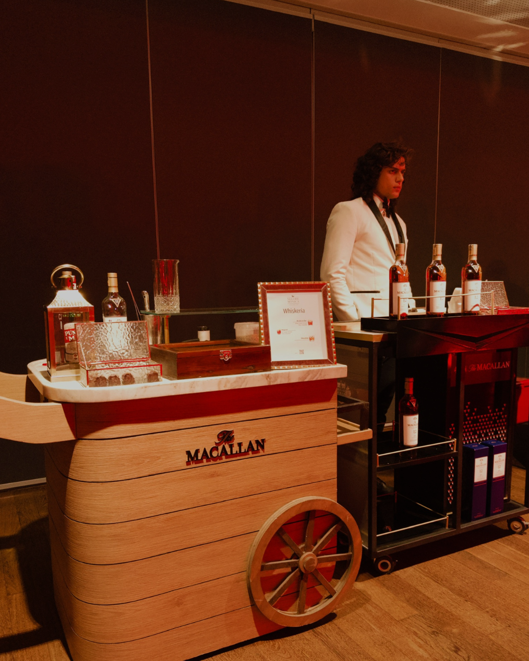 Balcão de coquetel com a marca Macallan e uma pessoa vestida de branco ao lado, com várias garrafas de uísque em exibição.