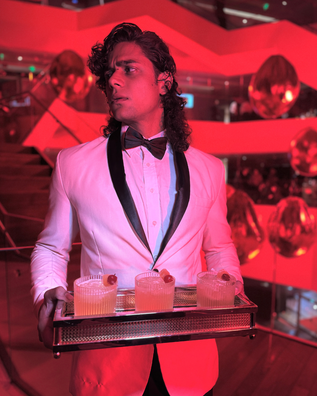 Homem vestindo terno branco com lapela preta, camisa branca e gravata borboleta, segurando uma bandeja com três copos de coquetel decorados com cereja em um ambiente com iluminação vermelha e decoração de bolas espelhadas.