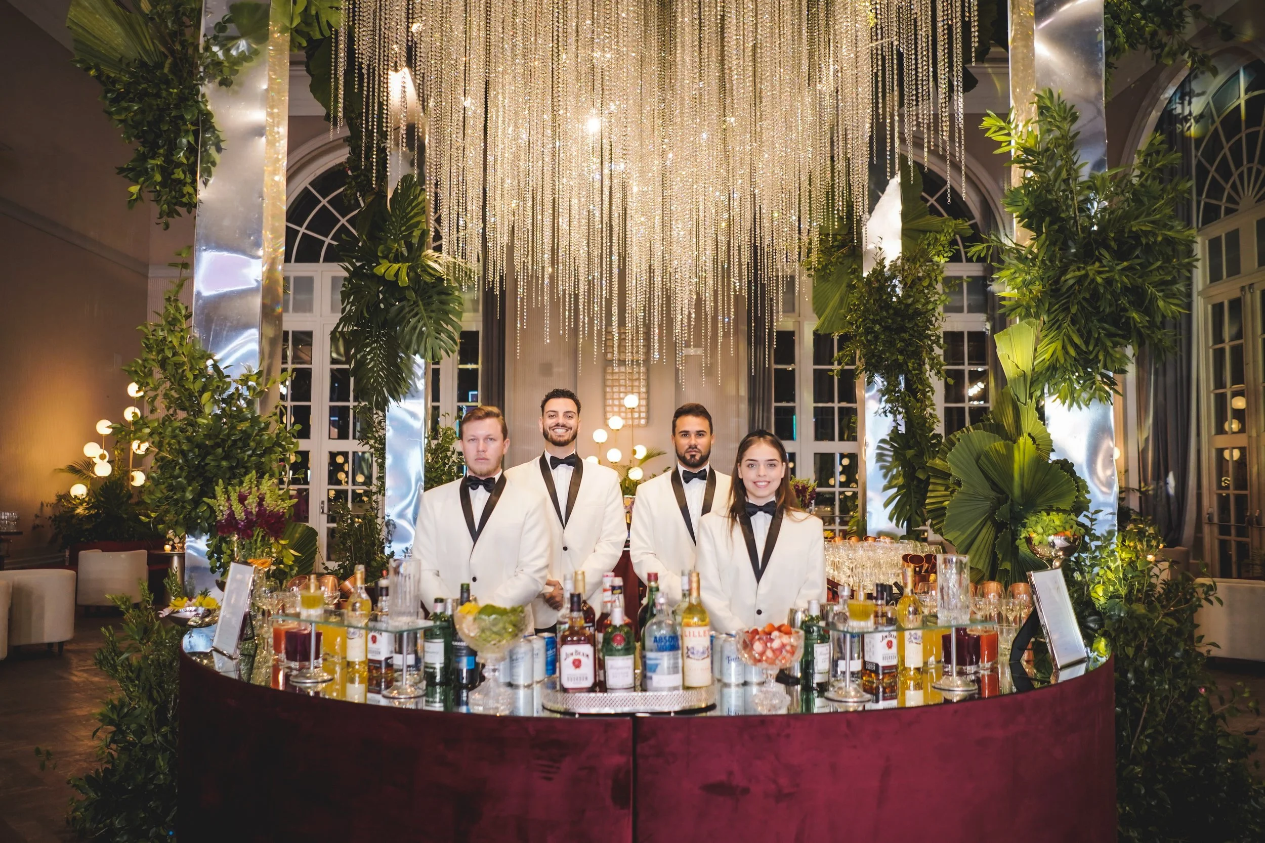 Equipe de quatro bartenders vestindo roupas brancas com gravatas pretas, posando atrás de um bar decorado com várias garrafas de bebidas e copos, em um salão com iluminação sofisticada, luzes penduradas e plantas verdes ao redor.