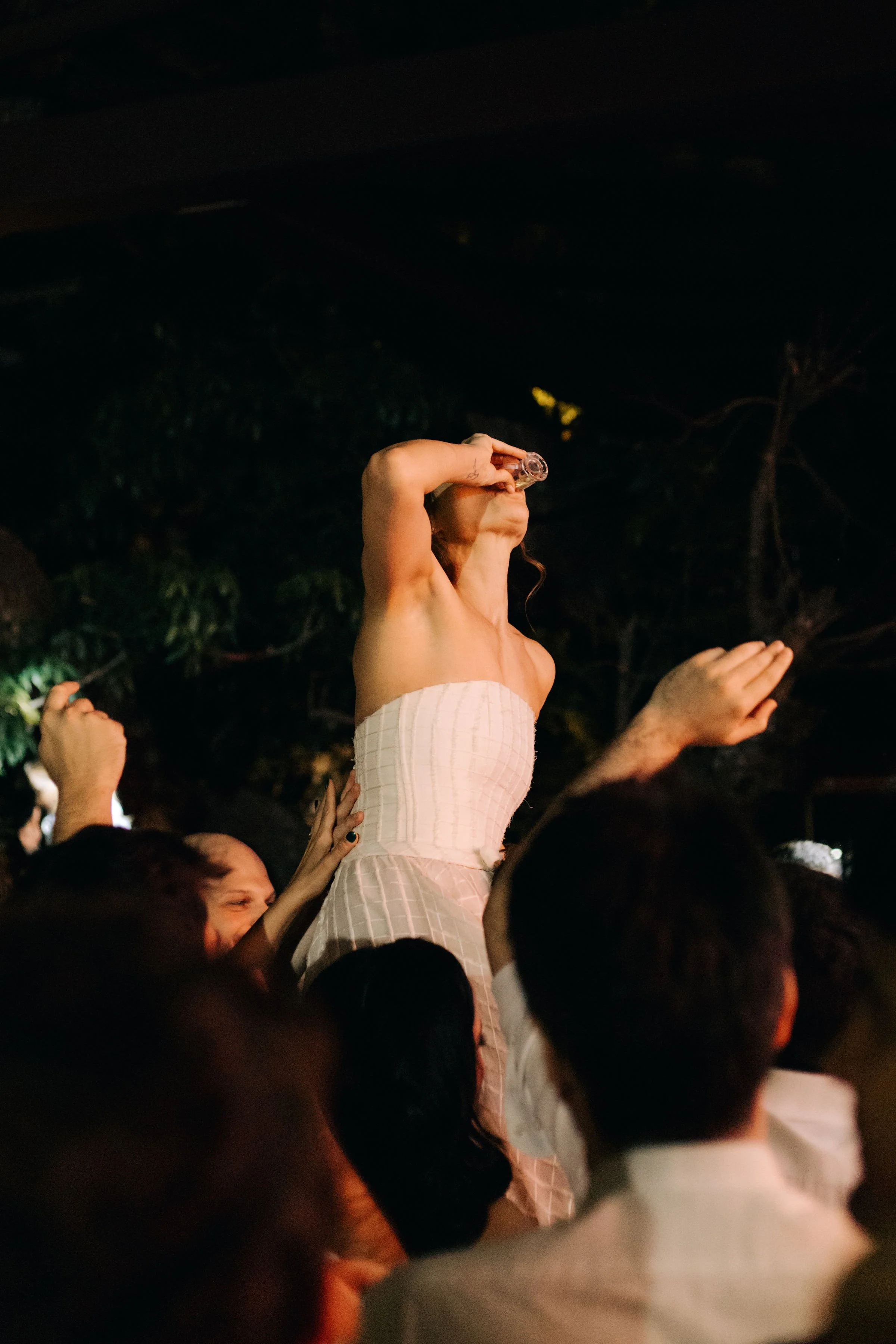 Mulher de vestido branco levantando uma taça durante uma celebração noturna rodeada por várias pessoas.