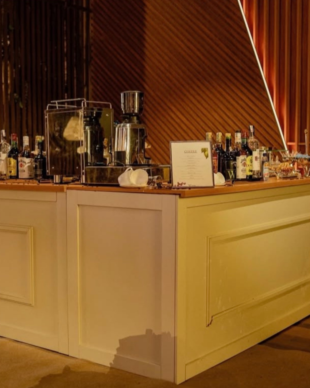 Bar com bebidas alcoólicas, máquina de café e utensílios, decorado com painel de madeira ao fundo.