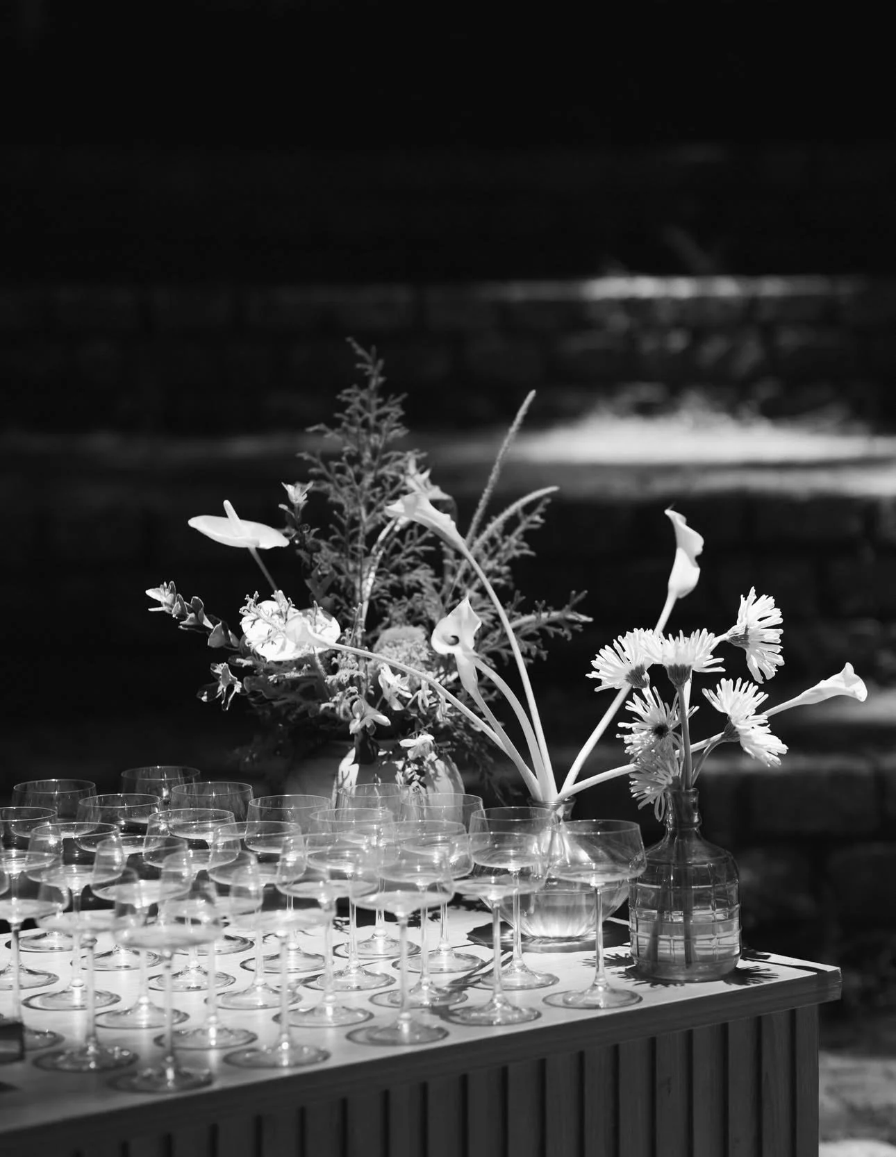 Mesa com várias taças de vidro e dois vasos com flores, uma com flores grandes e outra com flores menores, em fundo escuro.