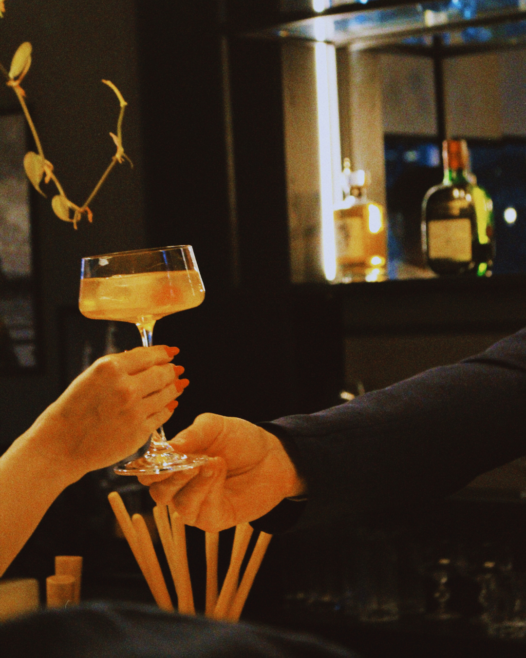 Pessoa entregando uma taça de coquetel para outra pessoa em um ambiente de bar ou restaurante à noite, com bebidas e decoração de fundo.