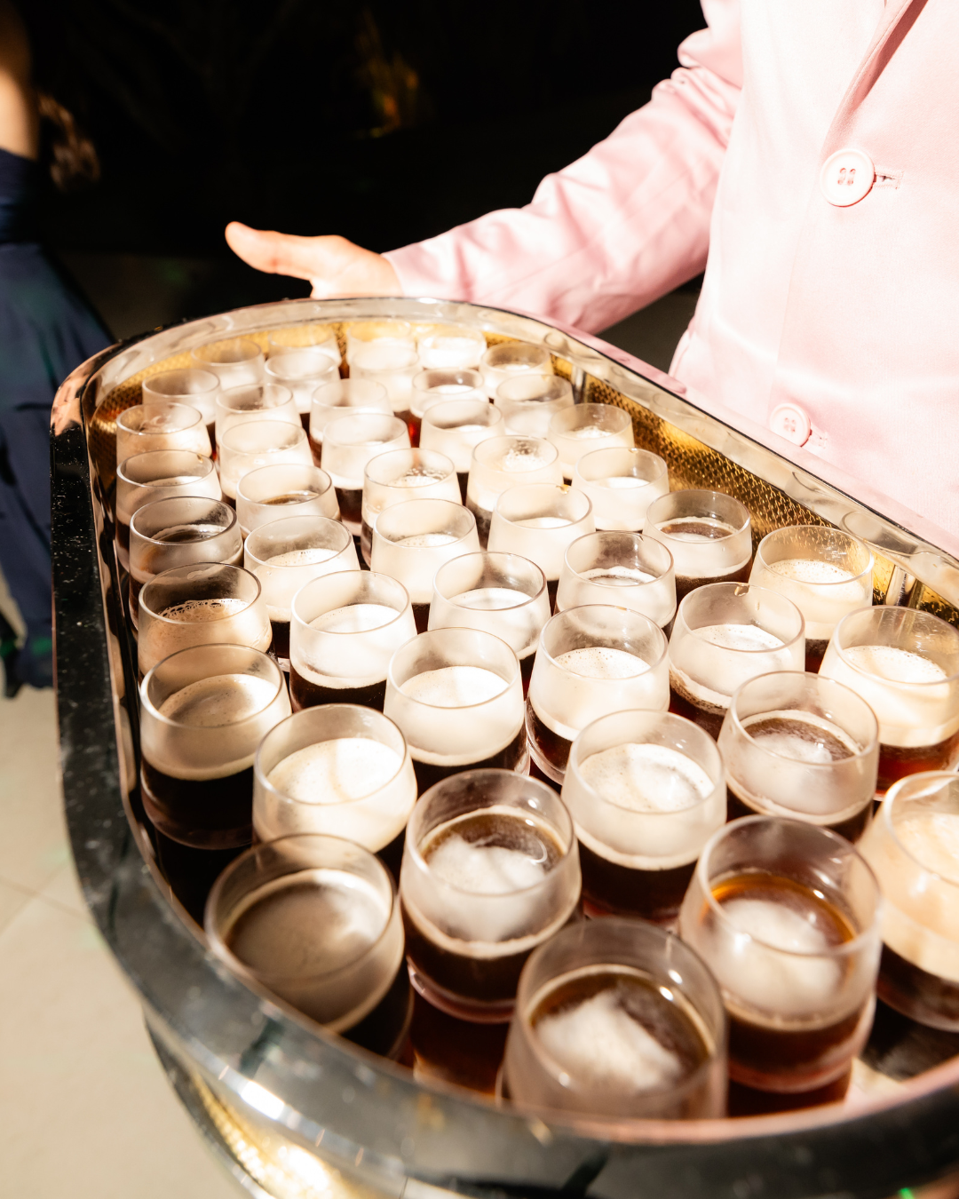 Pessoa servindo taças de vinho tinto em uma bandeja, em uma festa ou evento social, com uma pessoa usando paletó rosa ao fundo.