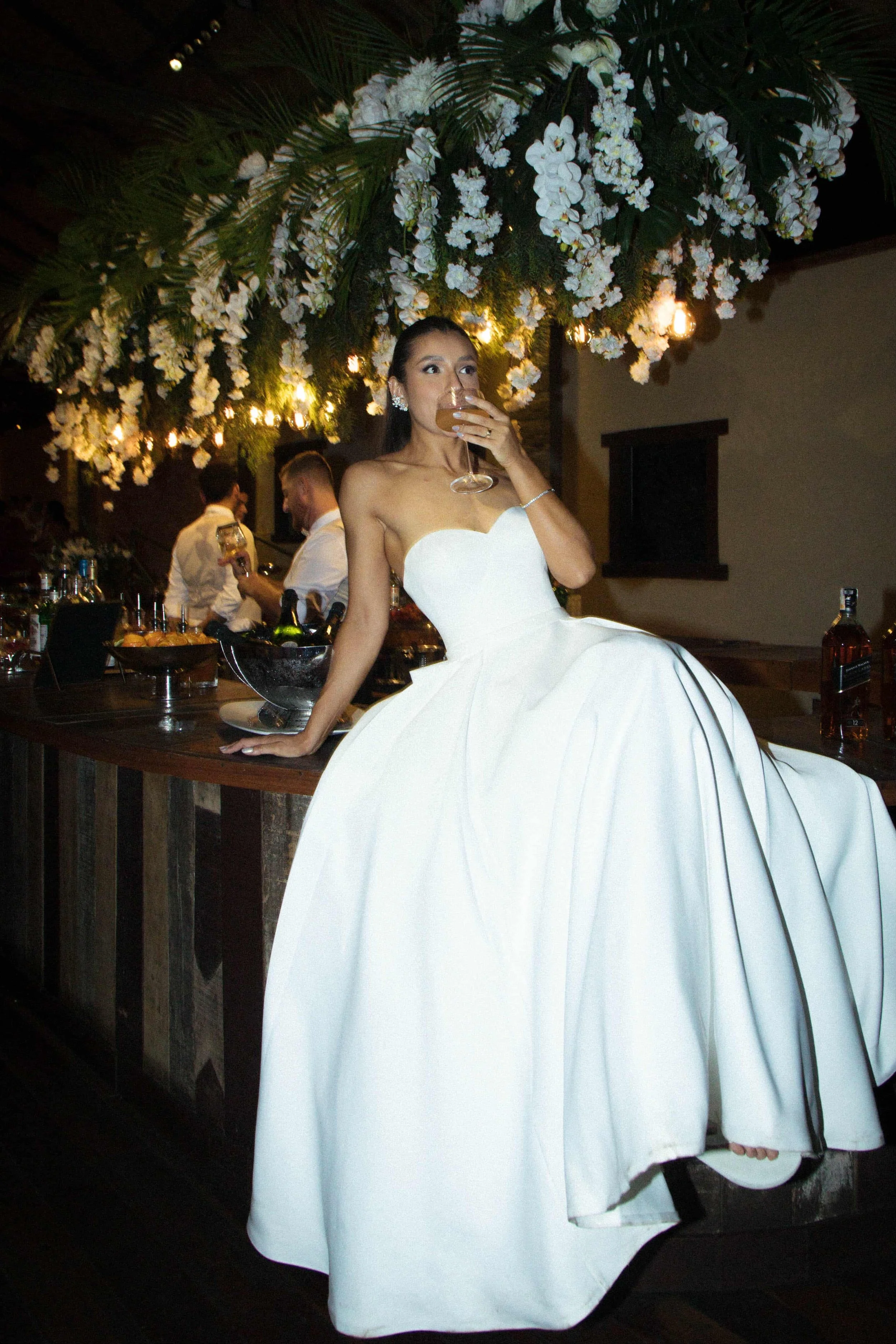 Noite de casamento, mulher em vestido branco longo, sentada em balcão decorado com flores brancas, segurando taça de vinho.