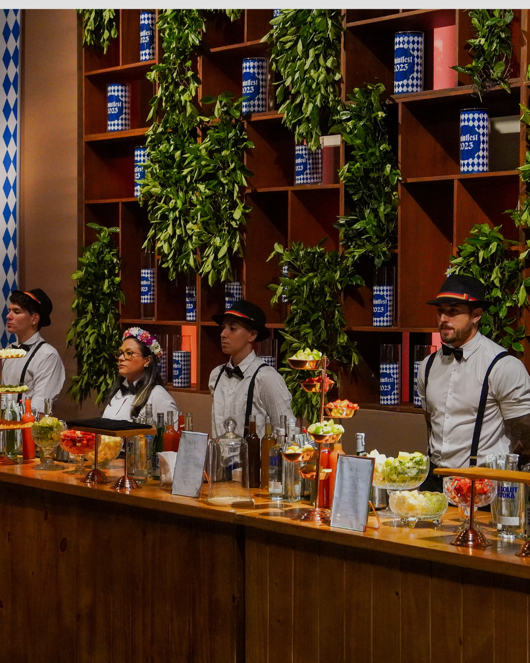 Pessoas vestidas com roupas de bartender olhando para o lado atrás de um balcão com bebidas e frutas, decorado com plantas verdes e prateleiras de madeira ao fundo.