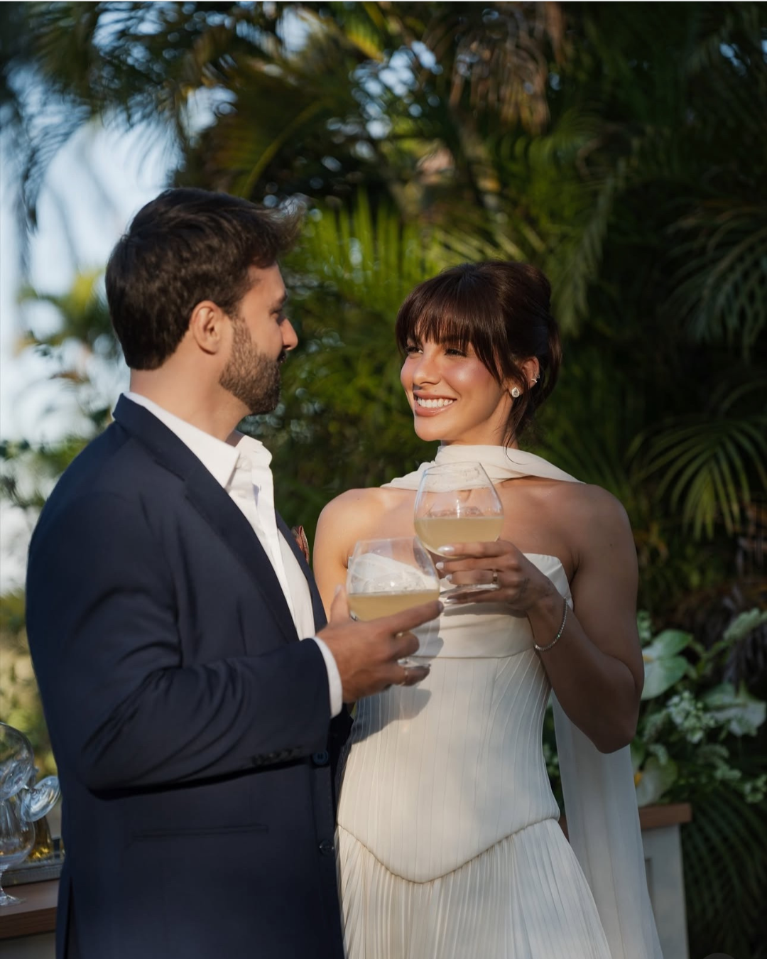 Casal sorridente em casamento, segurando taças de vinho branco, cercado por vegetação tropical.
