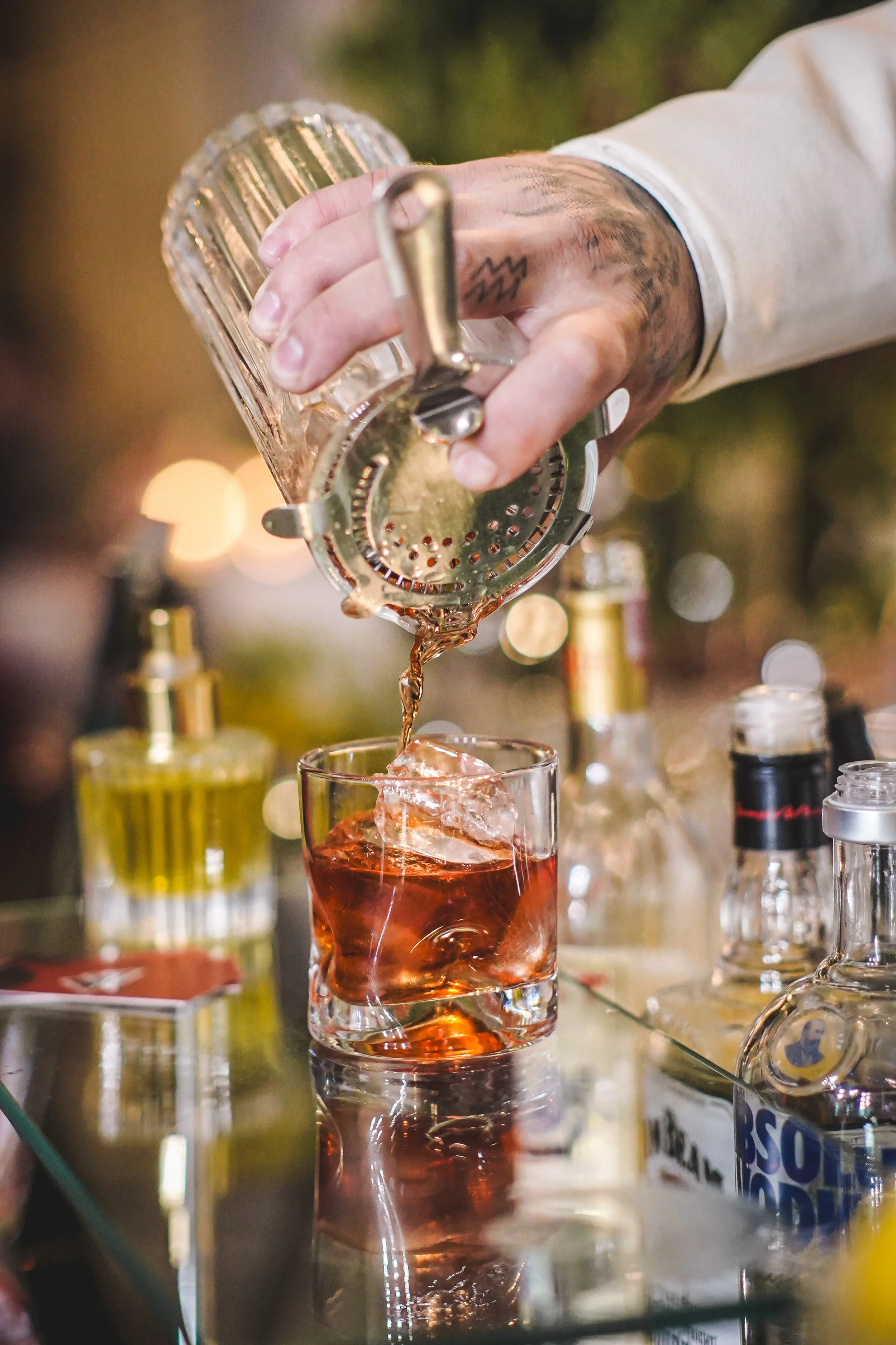 Pessoa servindo um drink alcoólico em um copo com gelo, usando uma coqueteleira de vidro. Várias garrafas de bebidas alcoólicas aparecem ao fundo.