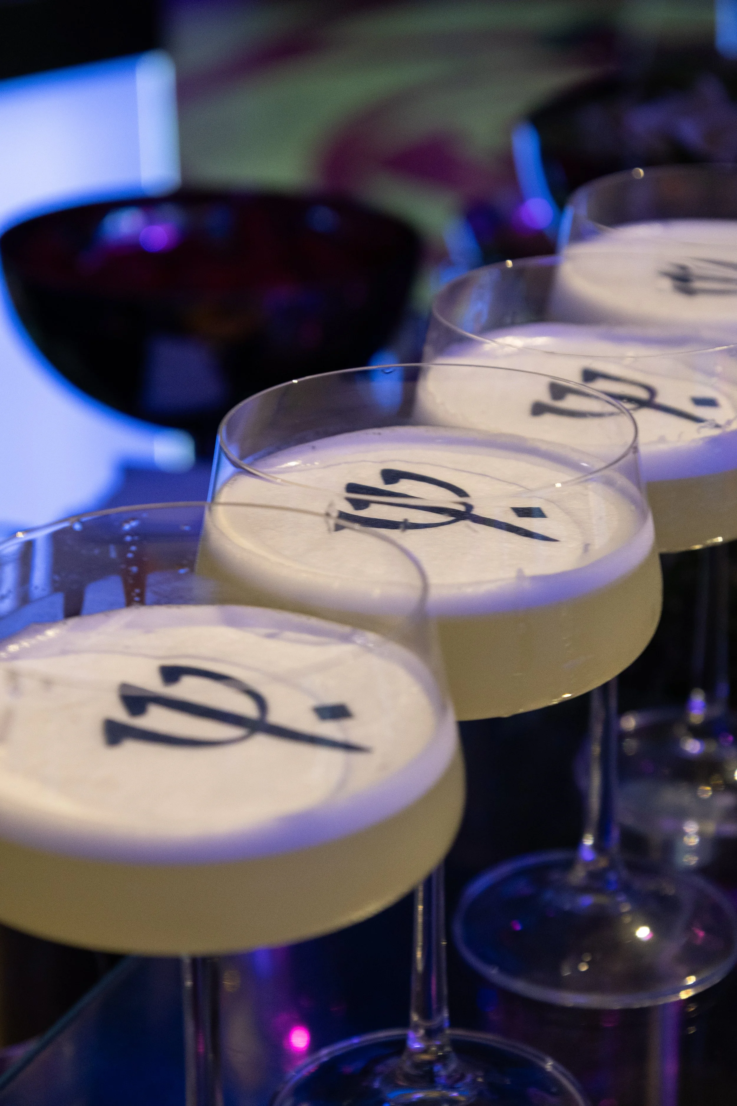 Copa de coquetel com espuma branca, decorada com letras pretas na parte superior, ao lado de uma taça de vinho tinto ao fundo.