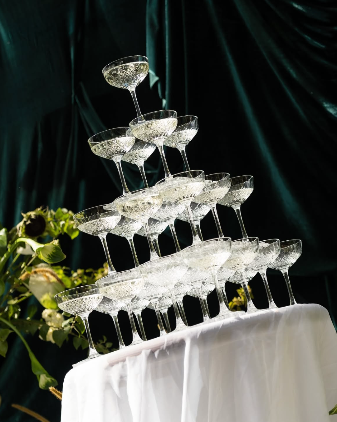 Taça de vidro decorada empilhada em formato de pirâmide sobre uma mesa com toalha branca, com fundo de cortina verde escura e flores verdes ao lado.