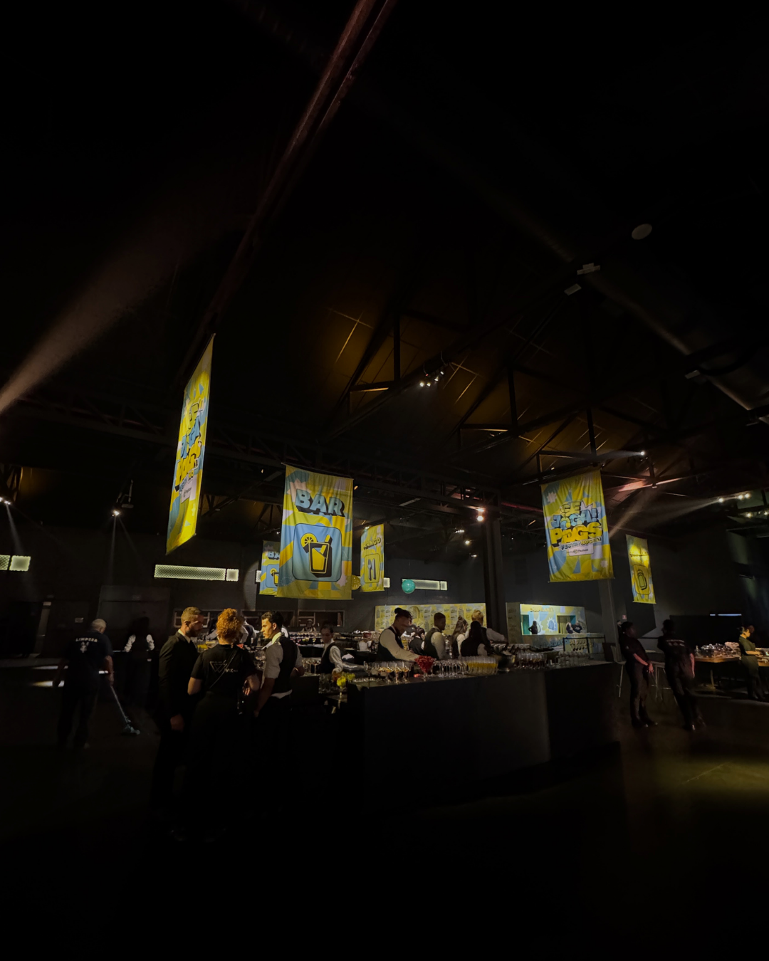 Bar com iluminação baixa, pessoas conversando e bartenders servindo bebidas, com banners coloridos no teto.