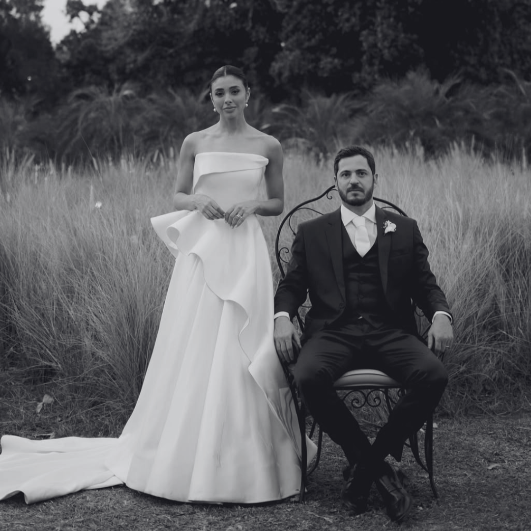 Casal de noivos posando ao ar livre, ela usando vestido de noiva branco e ele de terno preto, ele sentado em uma cadeira de ferro, ela de pé ao lado dele, ao fundo há grama alta e árvores.