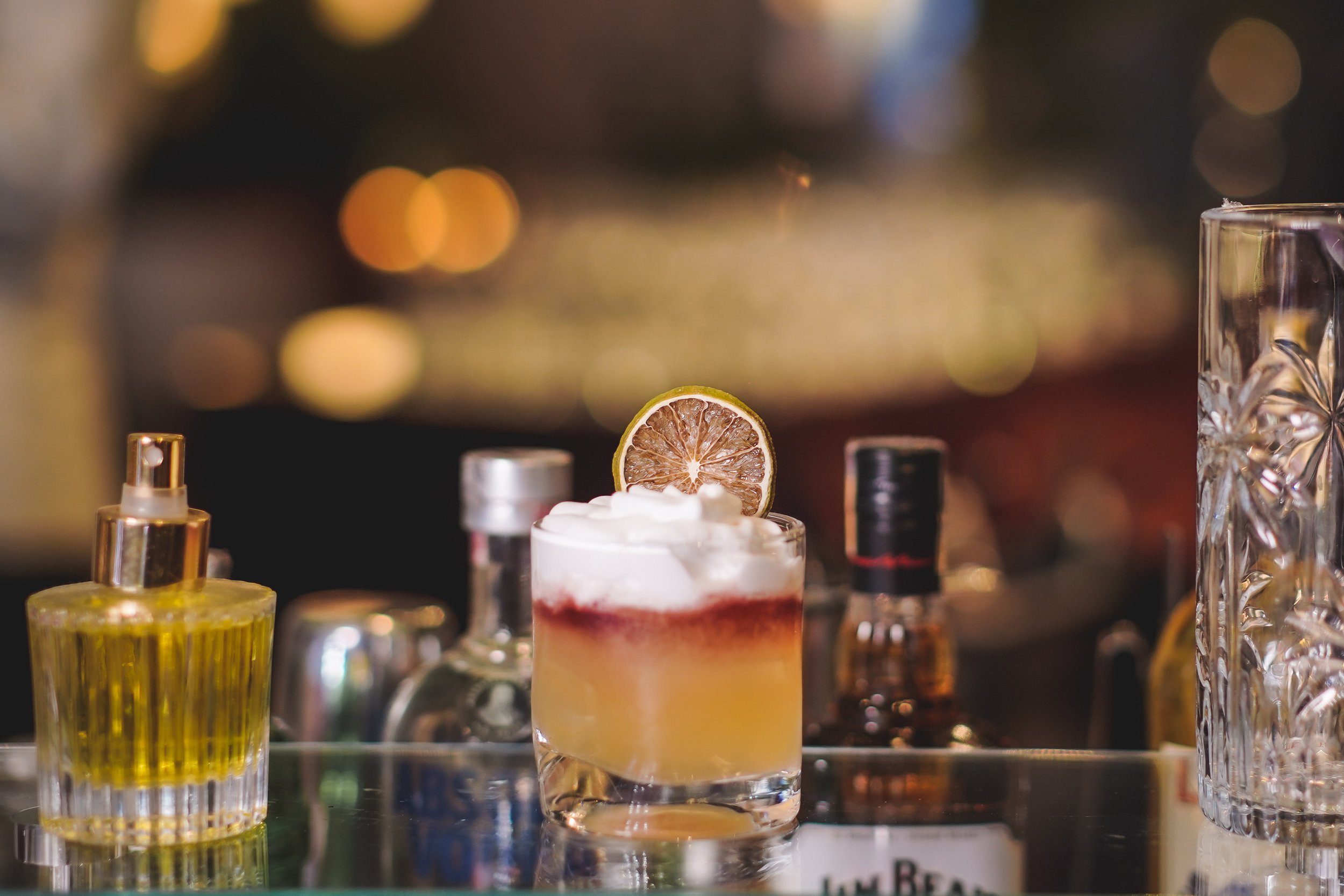 Copo de coquetel com limão, gelo, espuma branca e líquido amarelo, cercado por garrafas e frascos de ingredientes para drinks em um balcão de bar.