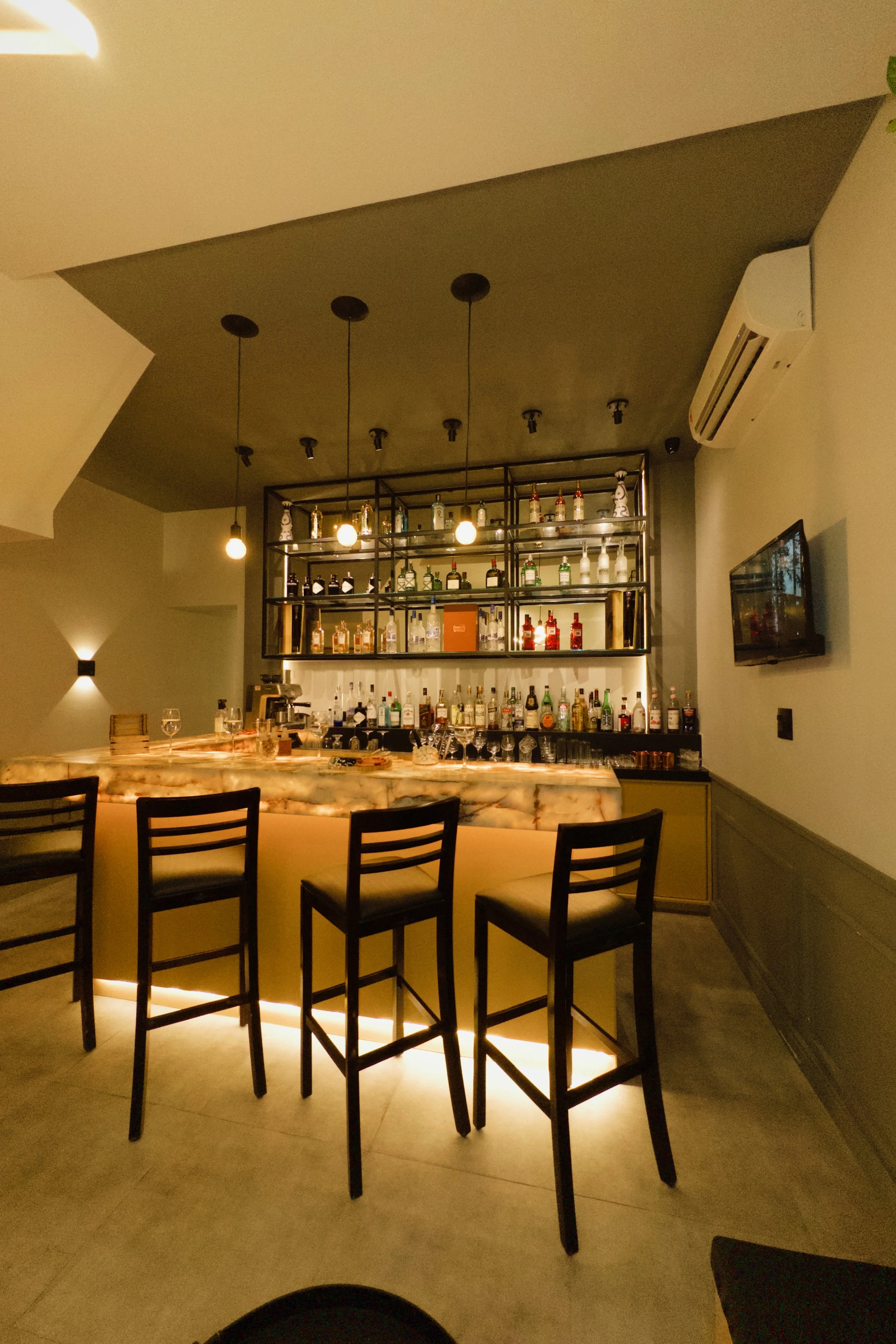 Bar com iluminação suave, estantes de bebidas com diversas garrafas, balcão de mármore iluminado e cadeiras de madeira na frente.