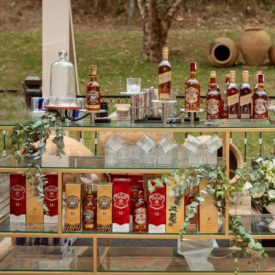 Bar de bebidas com várias garrafas de whisky Chivas Regal e acessórios, decorado com plantas e copos de vidro em um ambiente ao ar livre.
