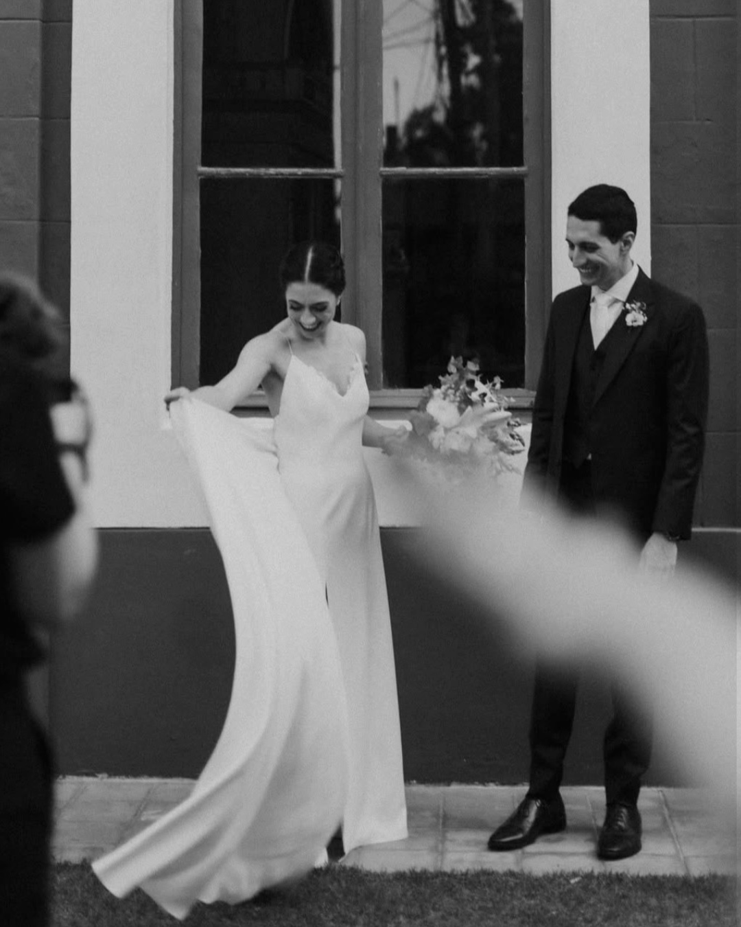 Recém-casados no casamento, mulher sorrindo e segurando um vestido, homem sorrindo com terno clássico, em ambiente interno com janela ao fundo.