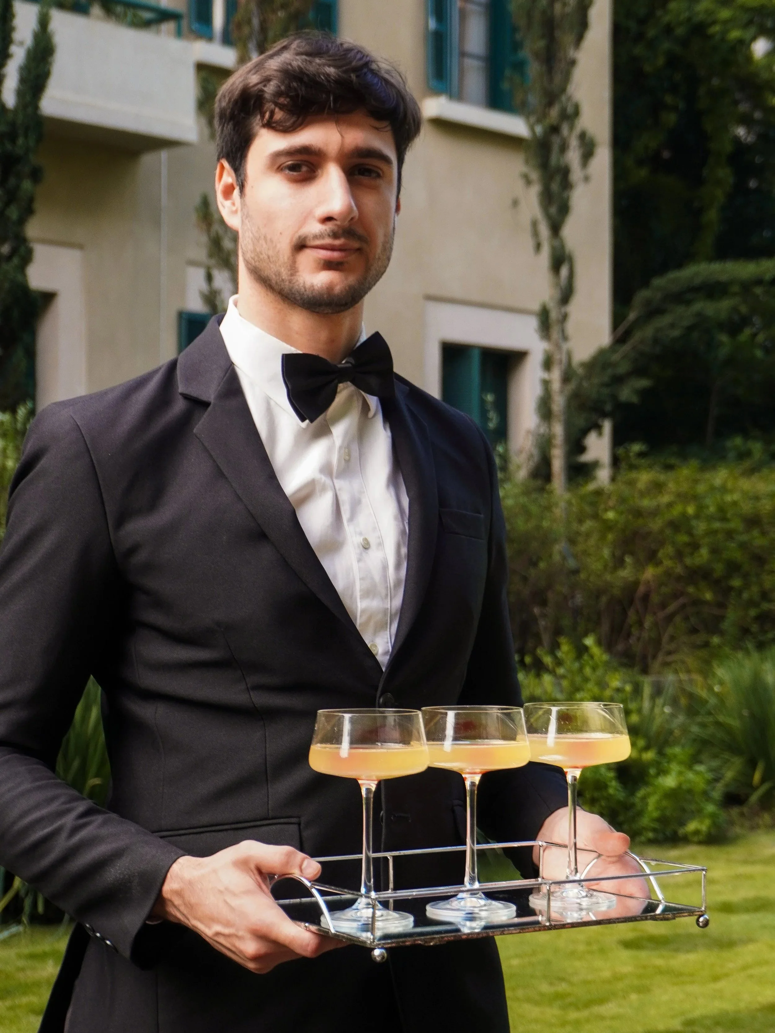 Homem elegante com terno preto, camisa branca e gravata borboleta, segurando uma bandeja com três taças de coquetel rosado, ao ar livre com jardim e edifício ao fundo.