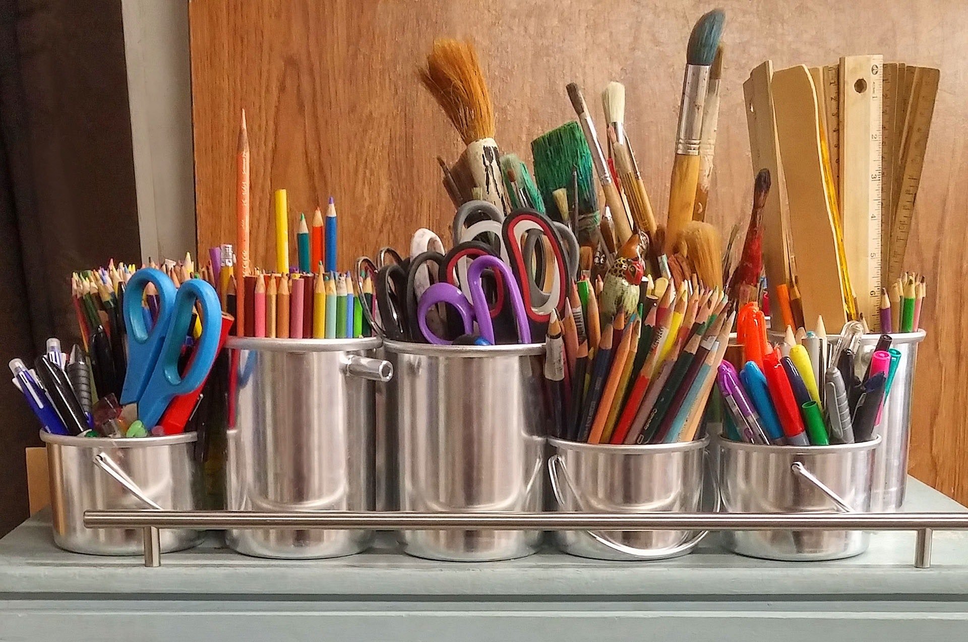 A metal organizer with multiple compartments holding various school and art supplies, including colored pencils, scissors, paintbrushes, rulers, and pens, on a table against a wooden wall.