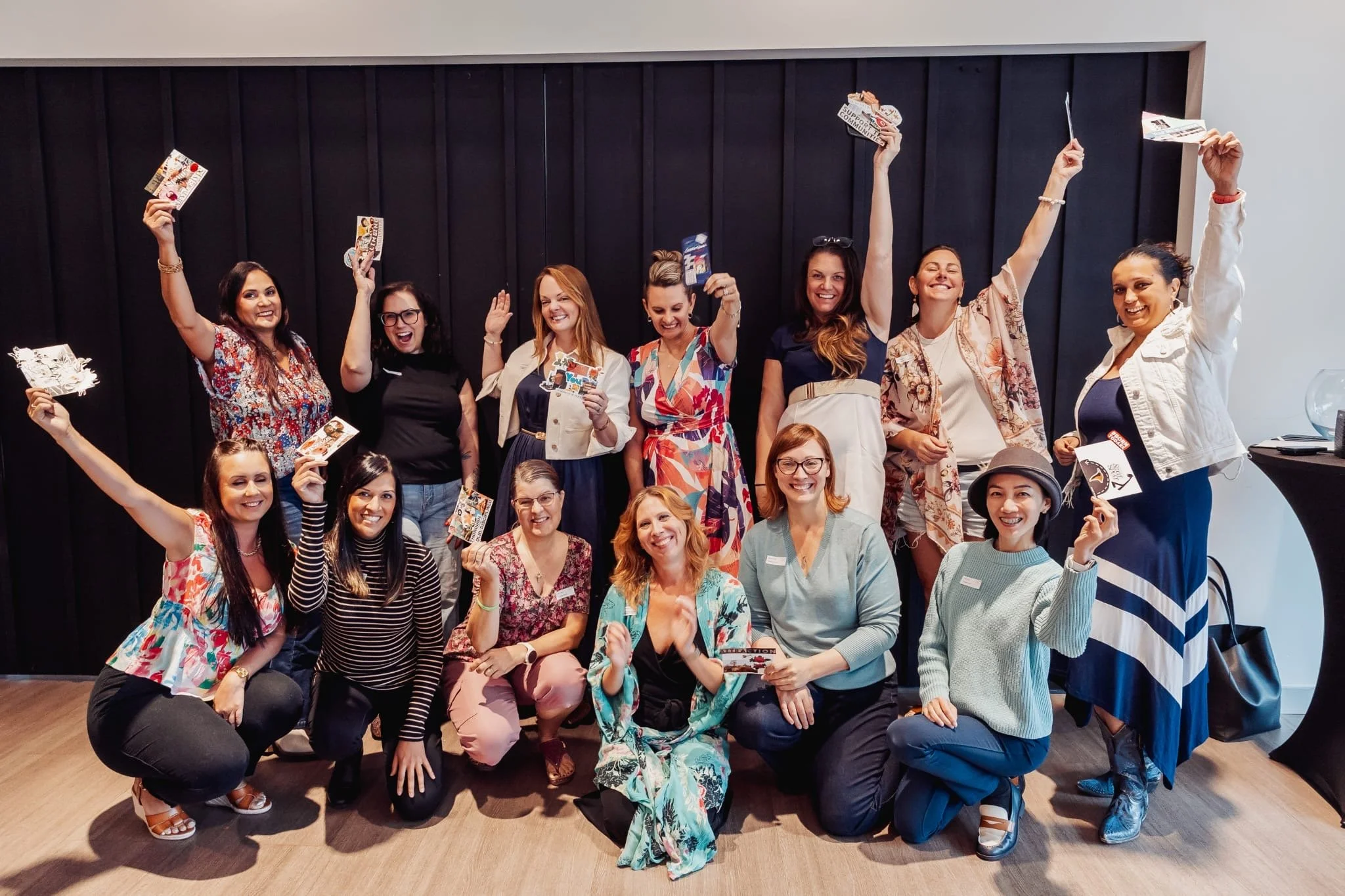 A group of fifteen women celebrating together, some standing and some kneeling, holding up paper items and smiling in front of a black wall.