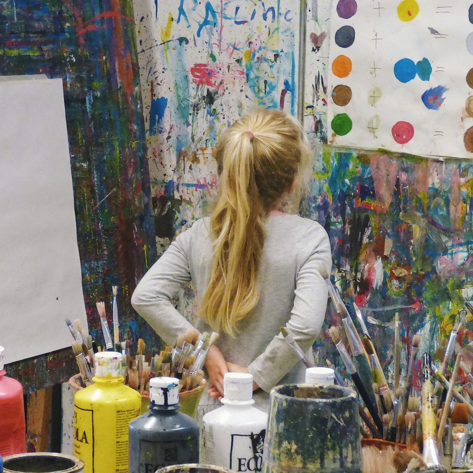 A young girl with long blonde hair, tied in a ponytail with a pink hair tie, stands in front of a paint-splattered wall and two large paintings or posters, looking at the artwork. She wears a light gray long-sleeve shirt. The surroundings include a table filled with paint bottles, brushes, and art supplies.