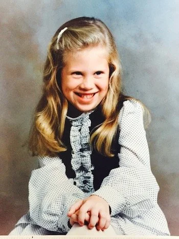 A young girl smiling with long blonde hair, wearing a white and black dress with a ruffled collar and a bow in her hair.