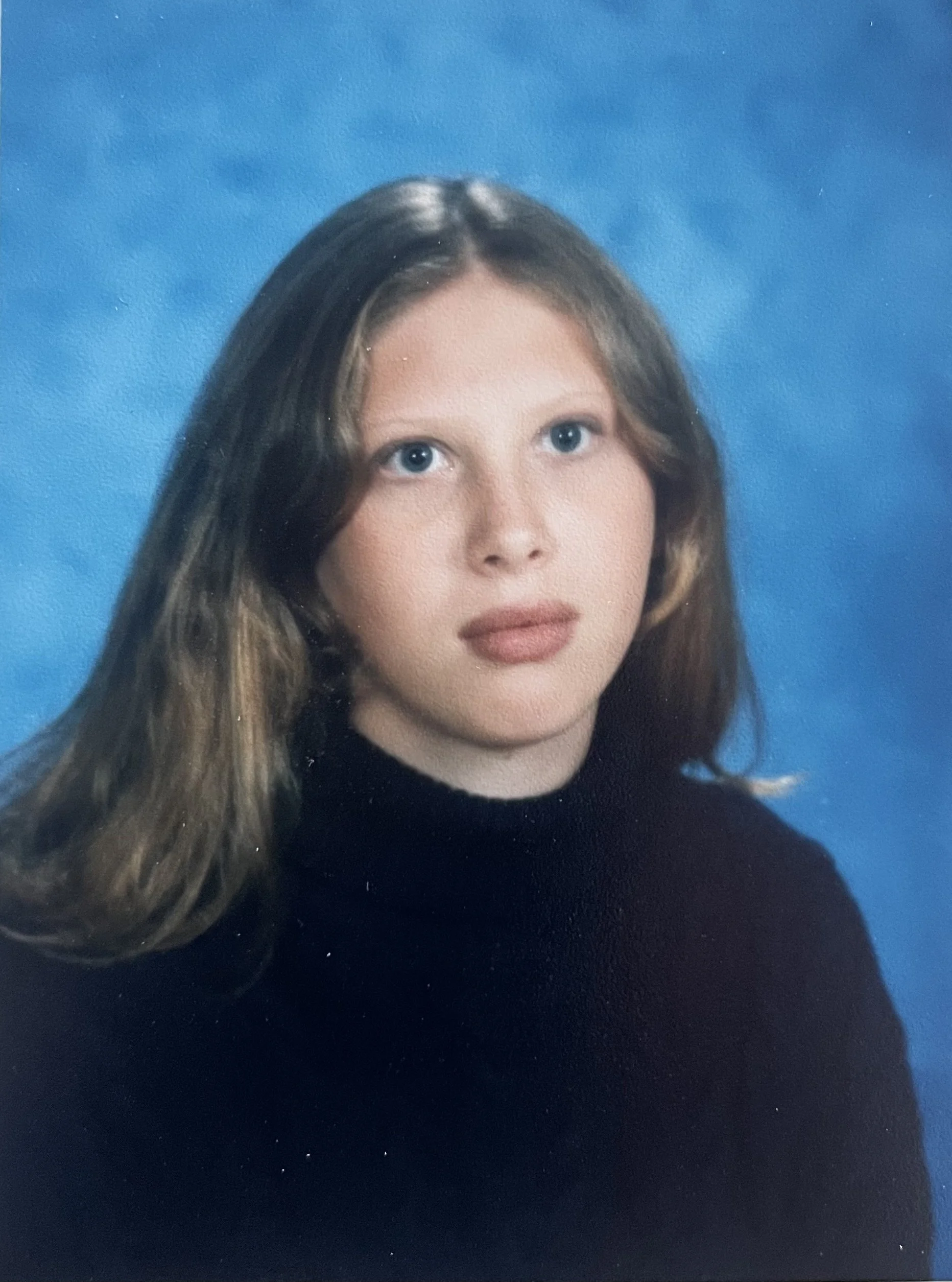 Studio portrait of a young woman with long brown hair, blue eyes, wearing a black turtleneck, against a blue background.