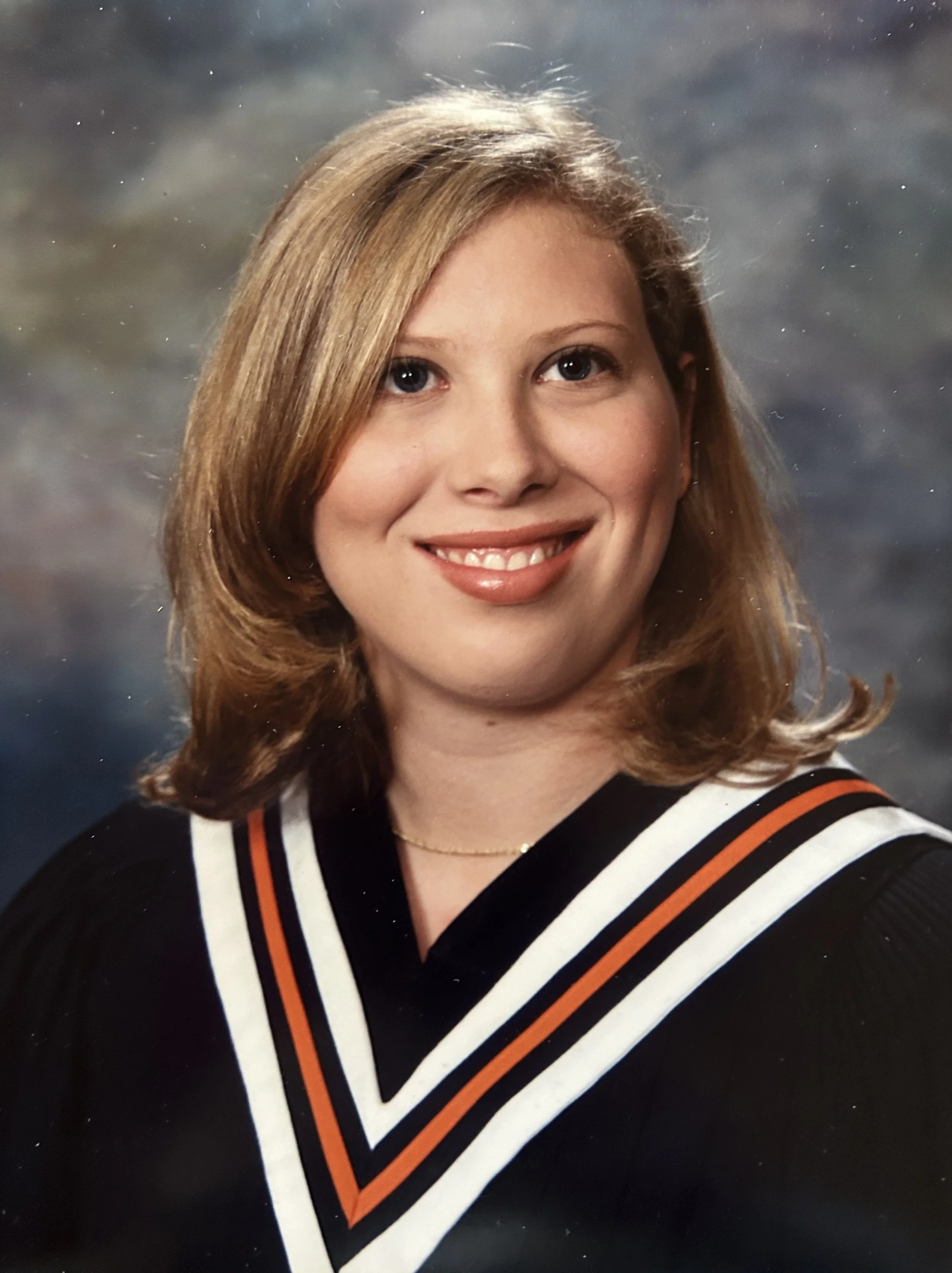 A woman with shoulder-length blonde hair wearing a black graduation gown with white, orange, and black trim, smiling in front of a dark, cloudy background.