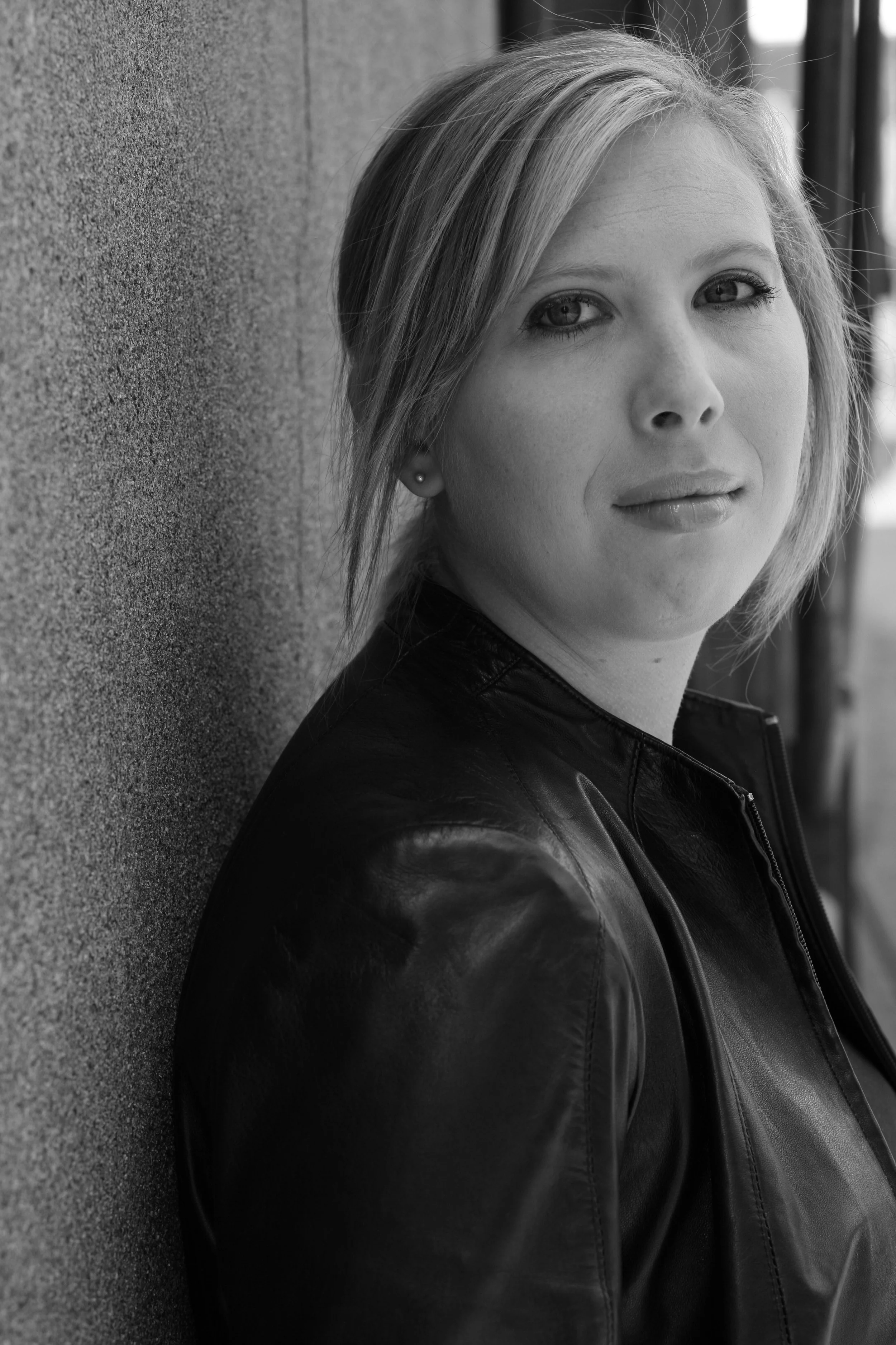 Black and white portrait of a woman with short blonde hair, wearing a leather jacket, leaning against a textured wall, looking at the camera.