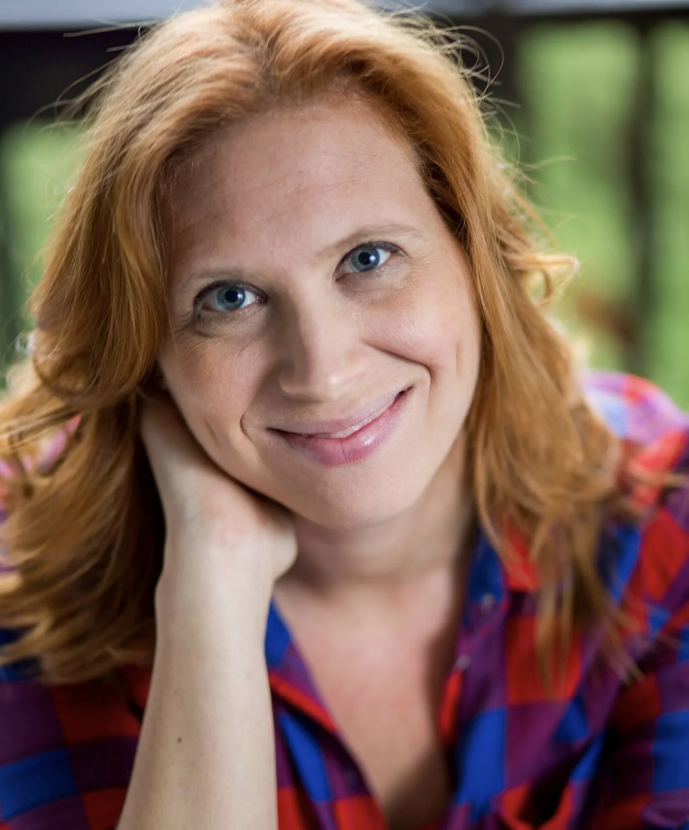 Close-up of a woman with red hair, smiling, resting her head on her hand, and wearing a colorful plaid shirt. Background is green and blurred.