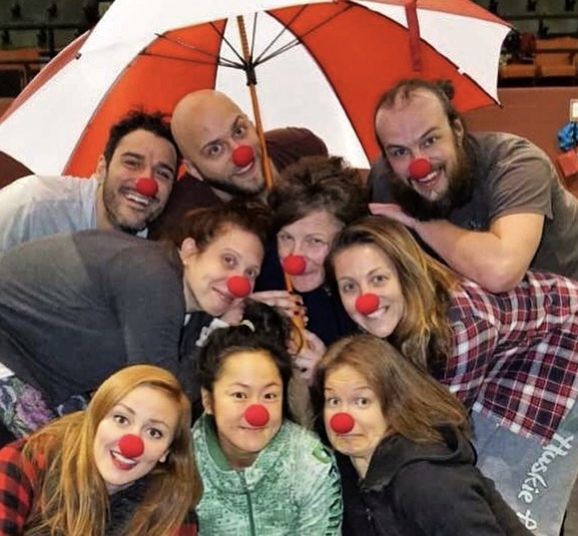 Group of people dressed as clowns with red noses, gathered under an umbrella, smiling and having fun.