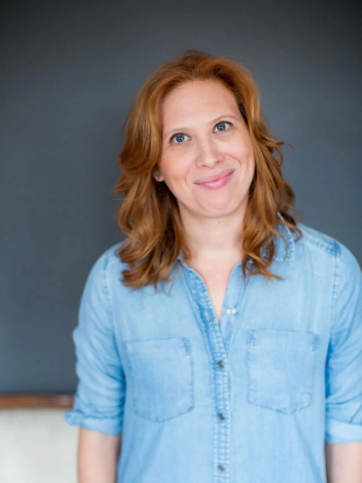 A woman with red hair, wearing a light blue denim shirt, smiling in front of a dark background.