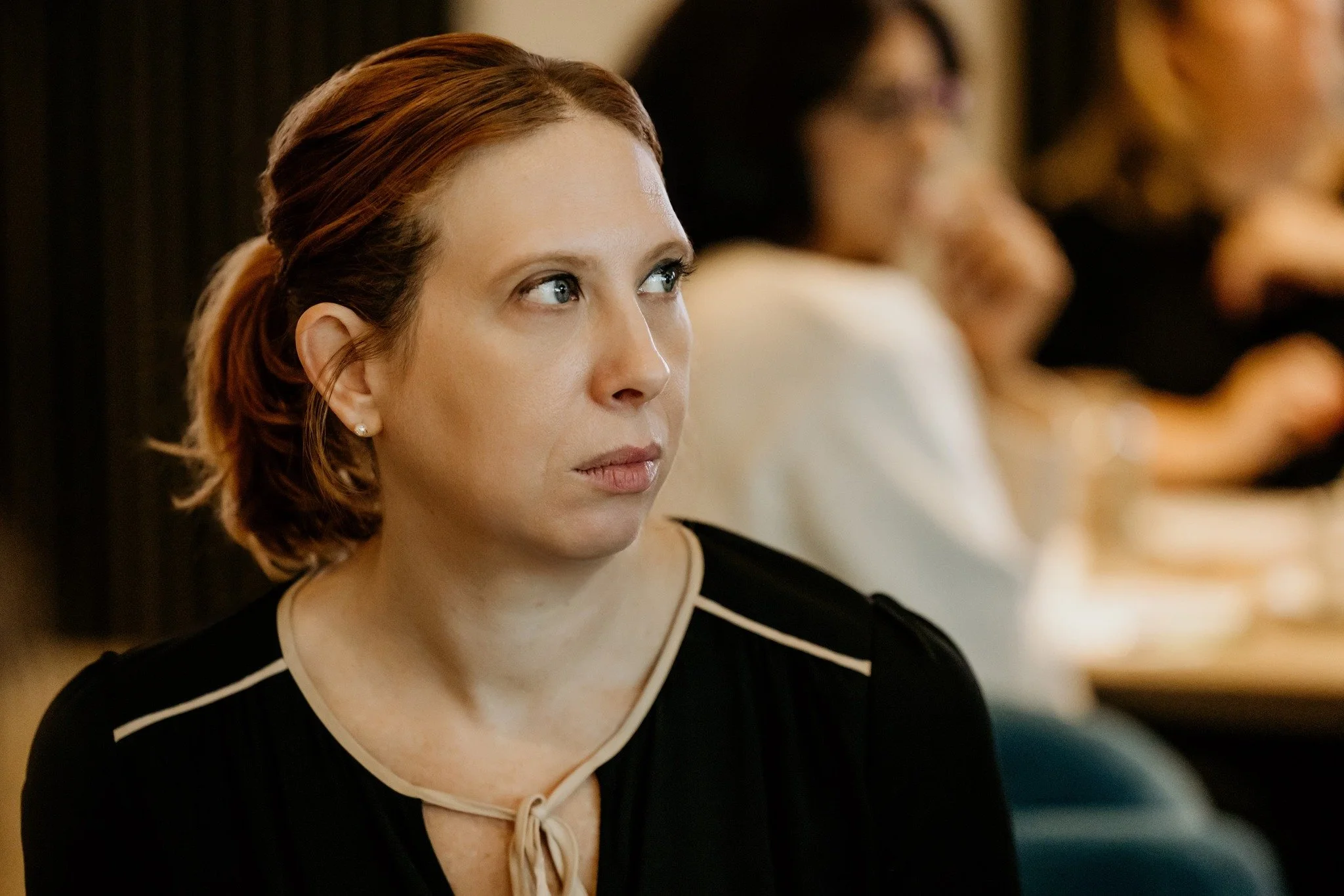 A woman with short, reddish-brown hair looking to her right in a room with blurred people in the background.