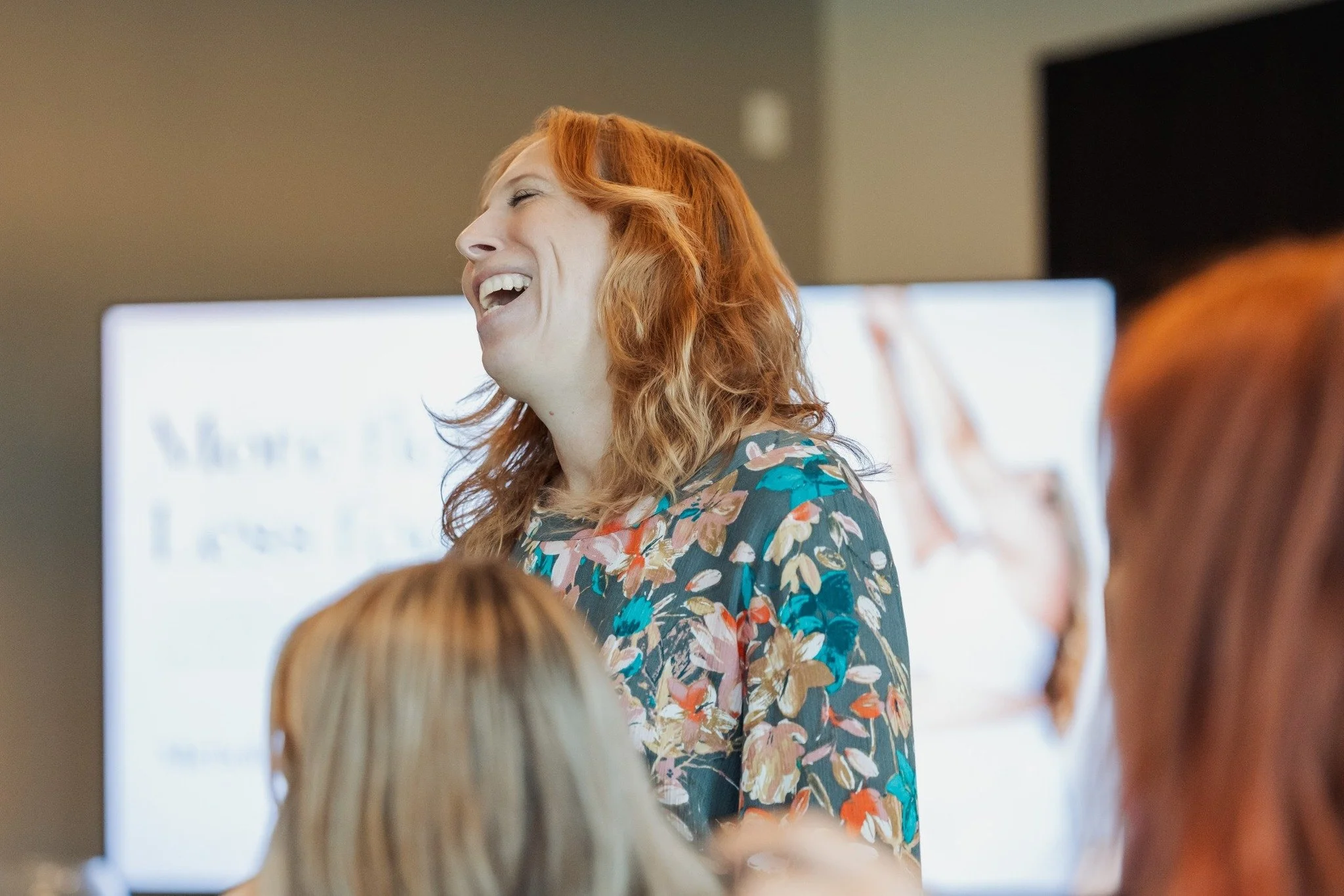 A woman with curly red hair and a floral blouse laughing in front of a group of people during a presentation or speech.