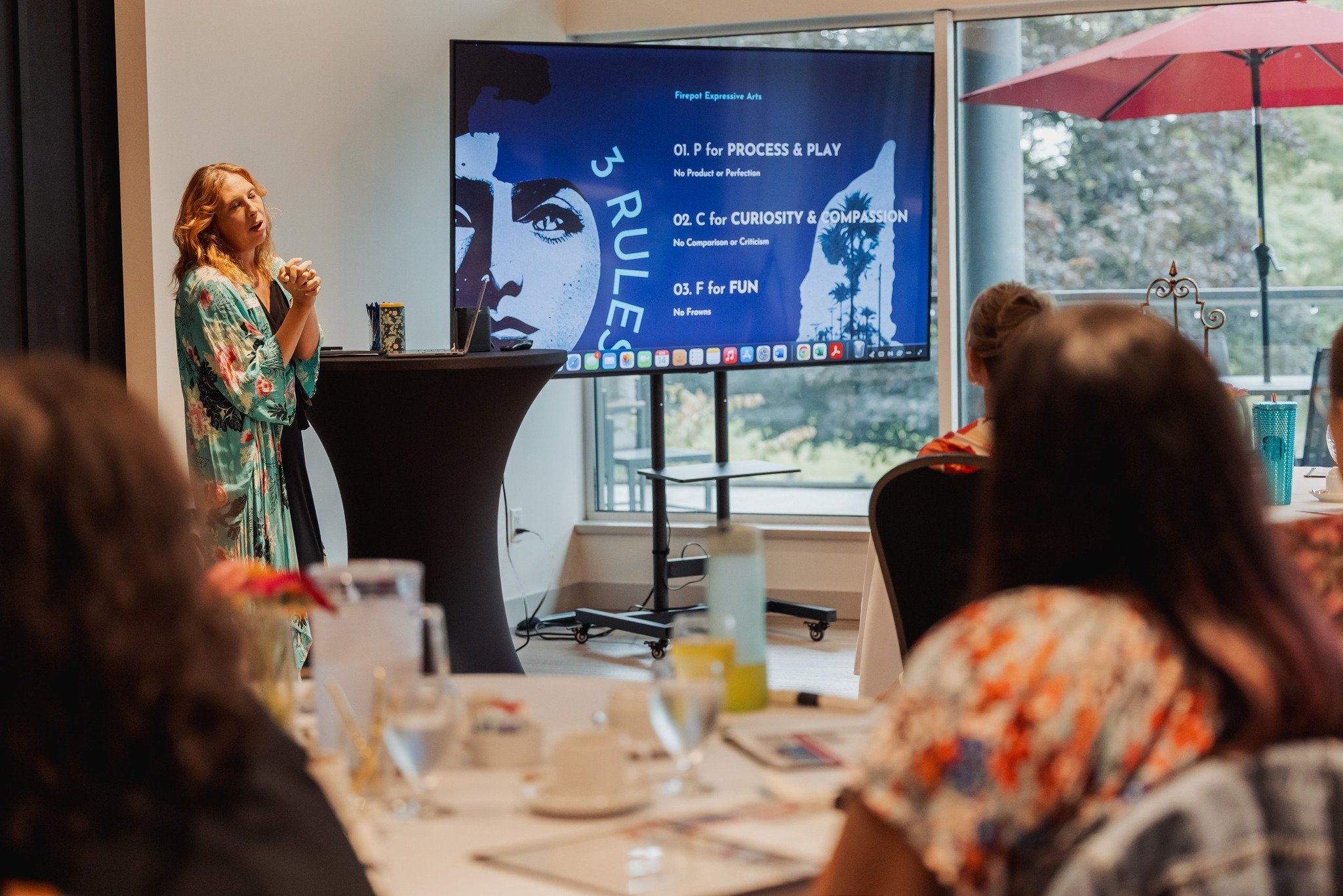 A woman giving a presentation in a conference room with large windows, a large screen displaying a list of rules, and an audience seated at tables.