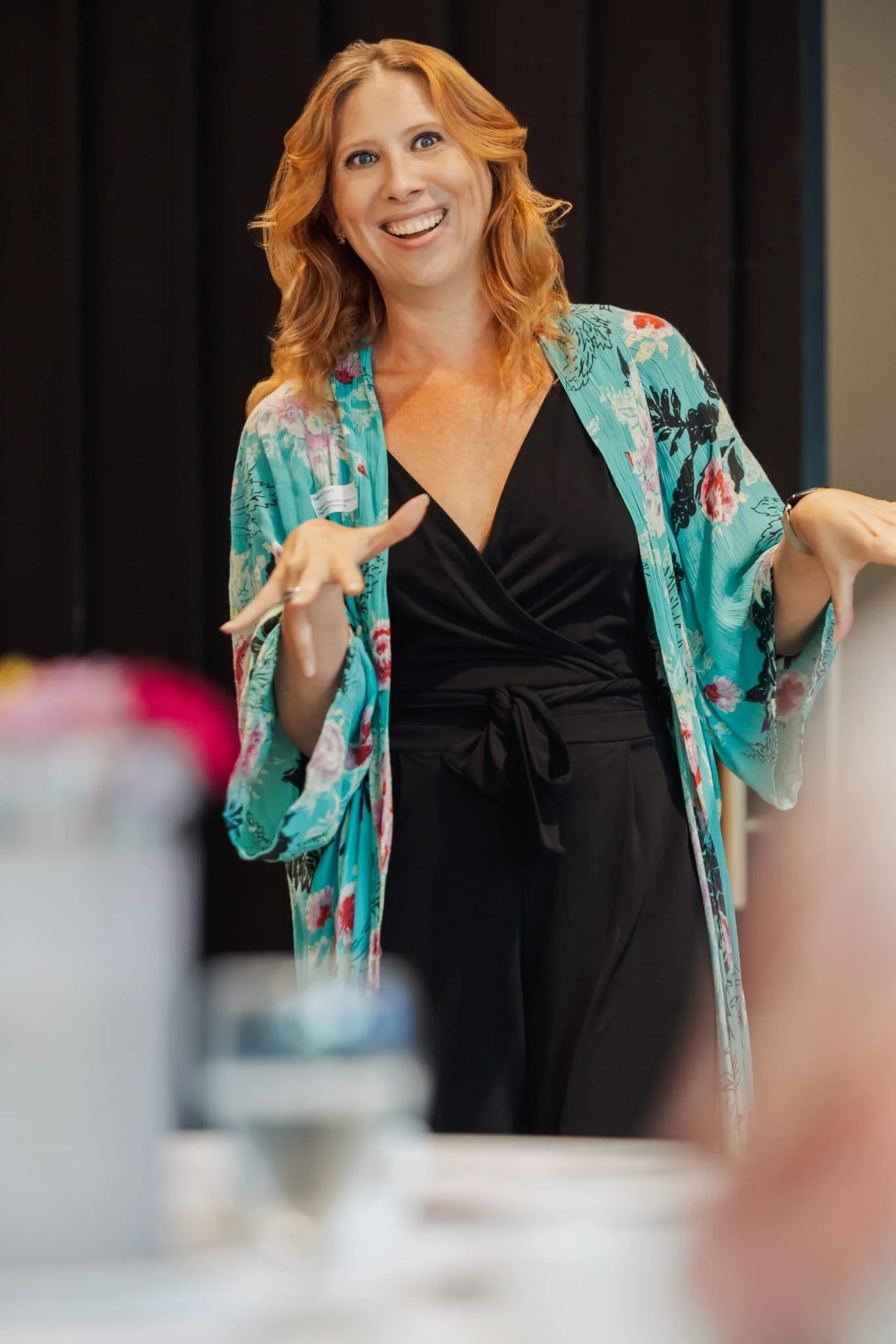A woman with red hair smiling and gesturing with her hands, wearing a black outfit and a floral kimono, standing in front of a dark background.