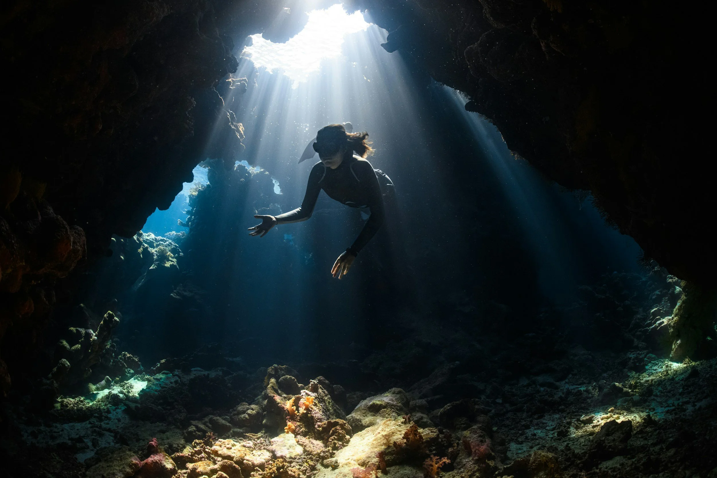 A person scuba diving underwater in a cave, with sunlight streaming through an opening above.